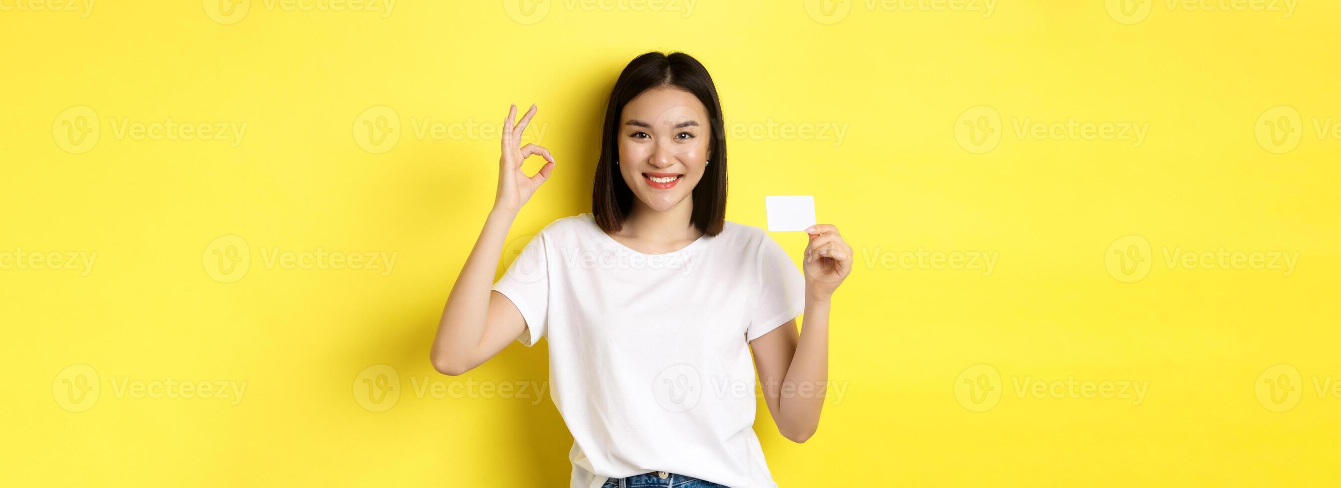 Young asian woman in casual white t-shirt showing plastic credit card and okay gesture, recommend bank, smiling at camera, yellow background photo