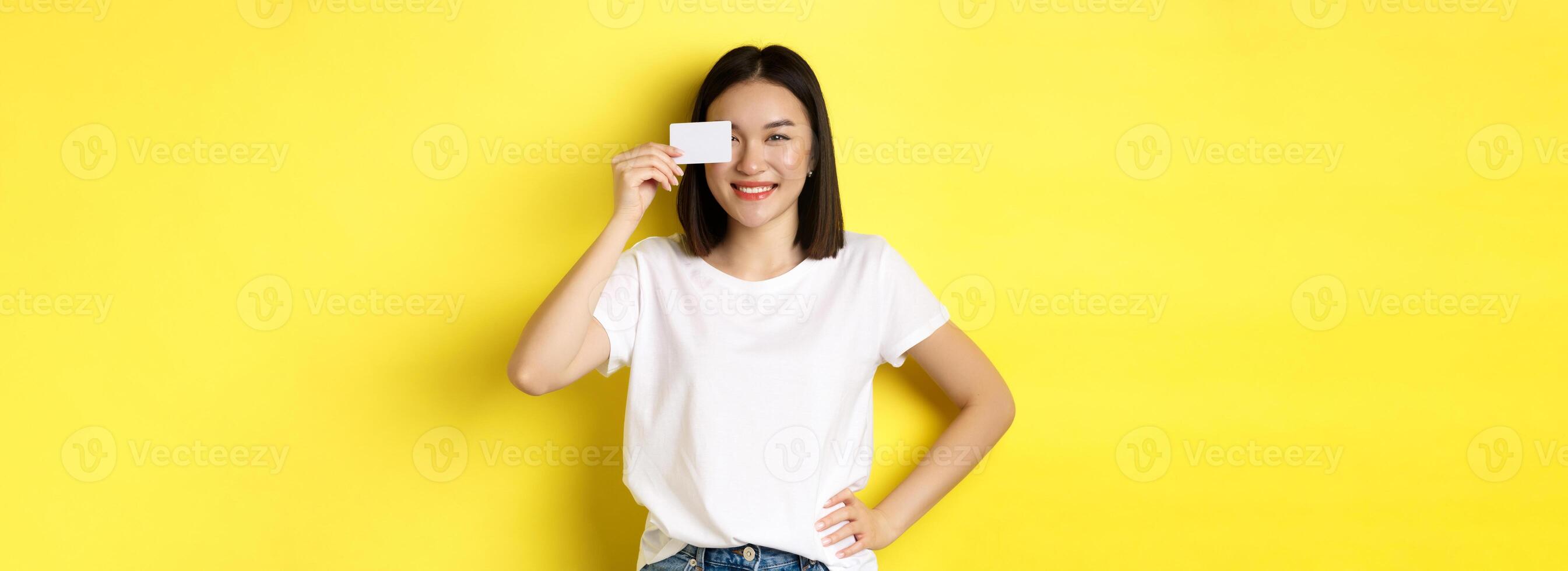 Young asian woman in casual white t-shirt showing plastic credit card and smiling at camera, yellow background photo