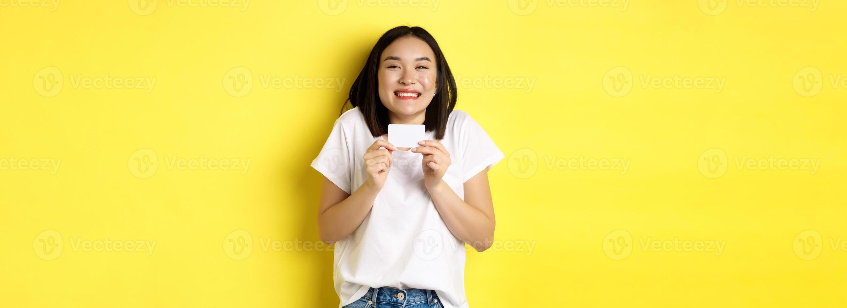 Young asian woman in casual white t-shirt going on shopping, showing plastic credit card and smiling excited, standing over yellow background photo