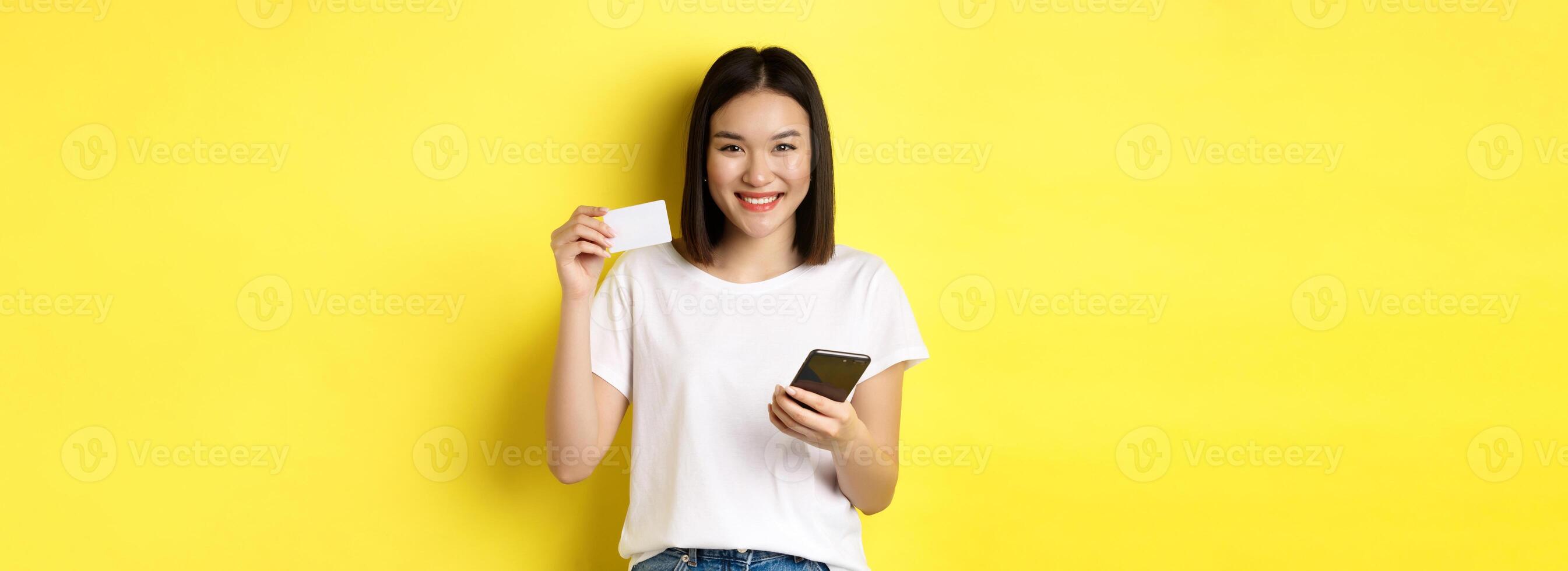 Attractive korean woman paying online with smartphone, showing plastic credit card and smiling, standing over yellow background photo