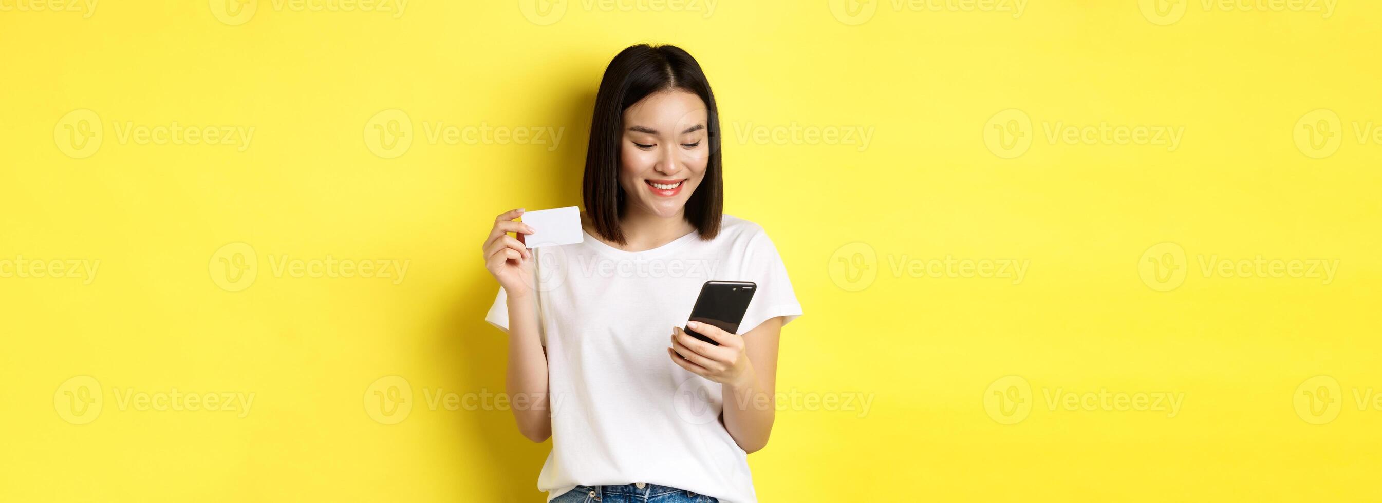 E-commerce and online shopping concept. Beautiful asian woman paying in internet, looking at smartphone screen and holding plastic credit card, yellow background photo