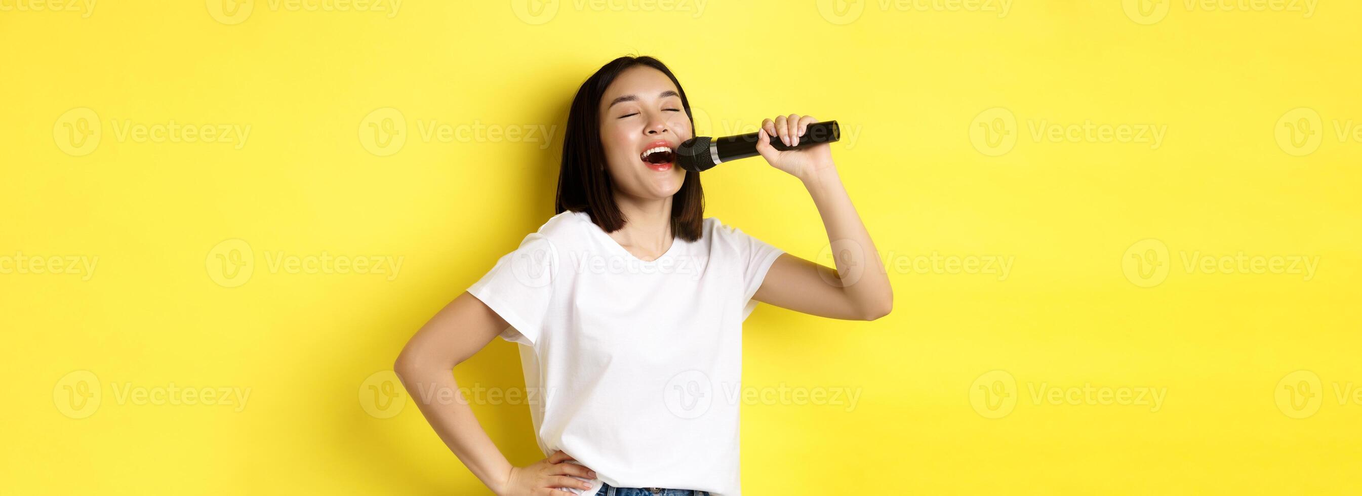 Happy asian woman singing song in karaoke, holding microphone, standing over yellow background photo
