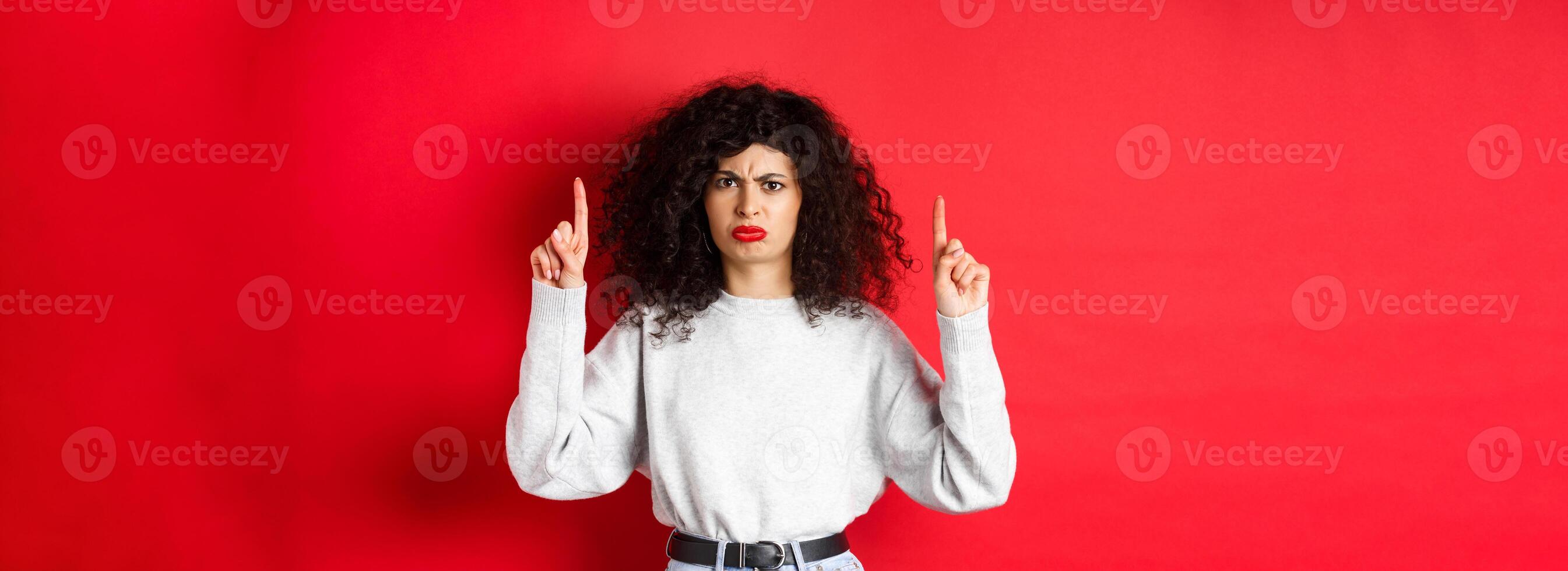 Grumpy young woman with curly hair frowning and grimacing unsatisfied, pointing fingers up at something bad, complaining on company, red background photo