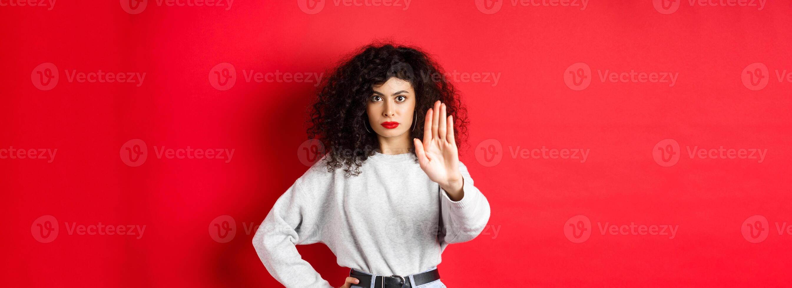 Confident tensed woman extend hand to say stop, disapprove action and prohibit it, make no gesture, standing on red background and forbid something photo