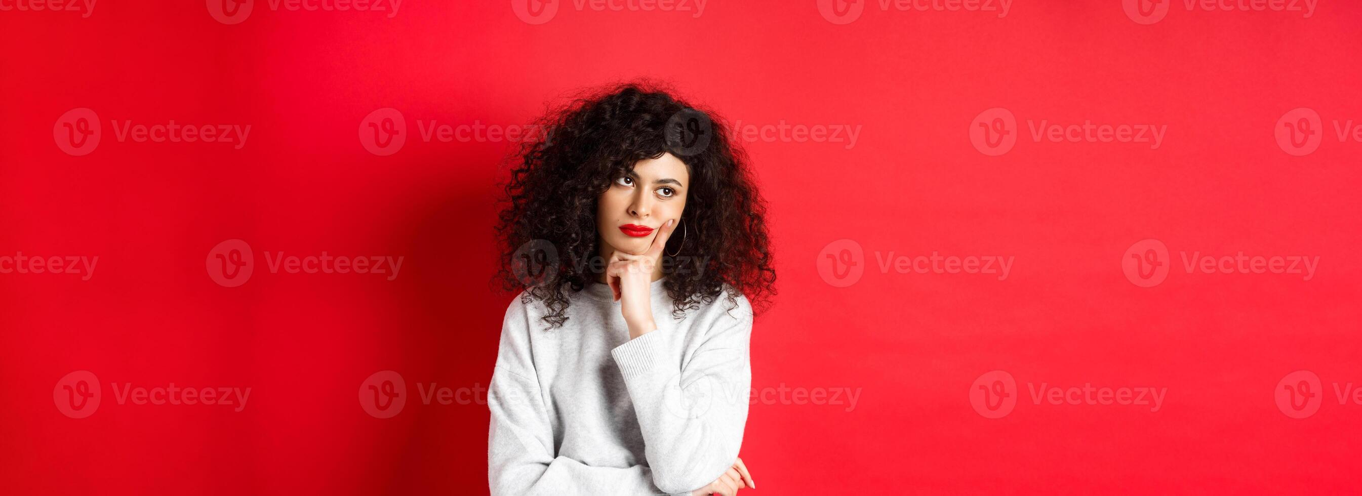 Grumpy young woman with curly hair, looking annoyed or bored at empty space, standing pensive and sad on red background photo