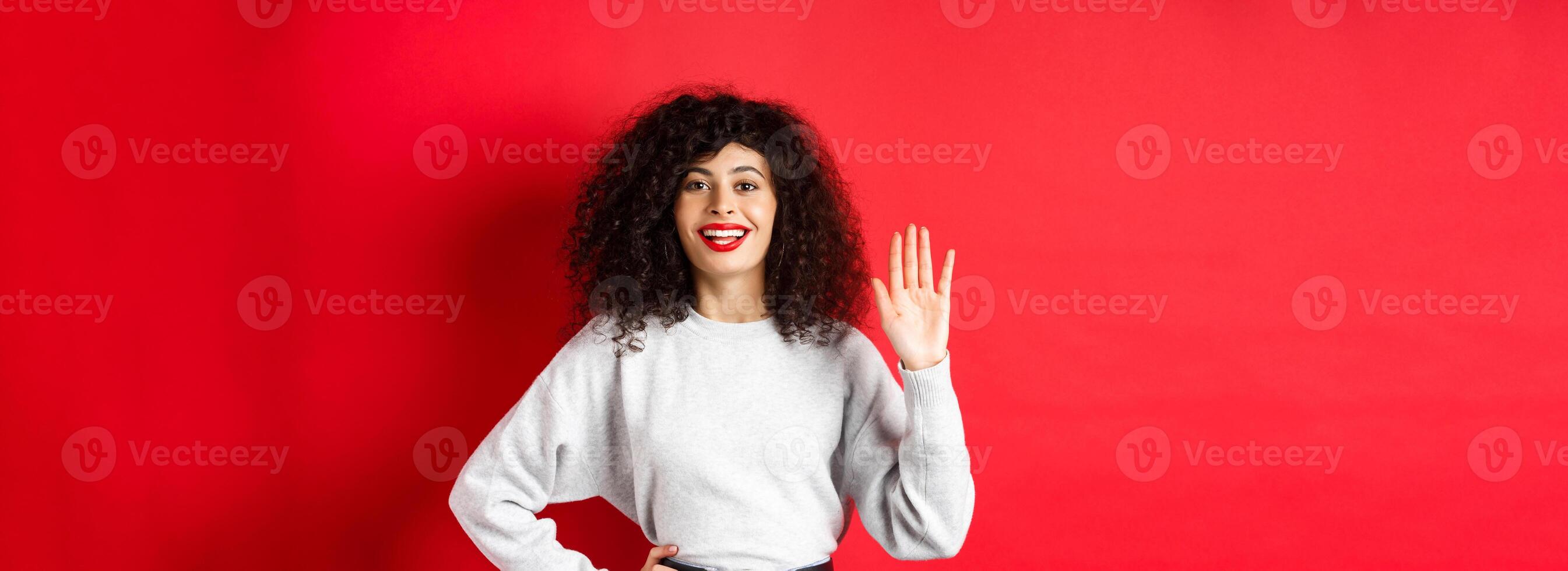 Friendly woman with curly hair greet you, waiving hand and saying hello, introduce herself, standing on red background photo