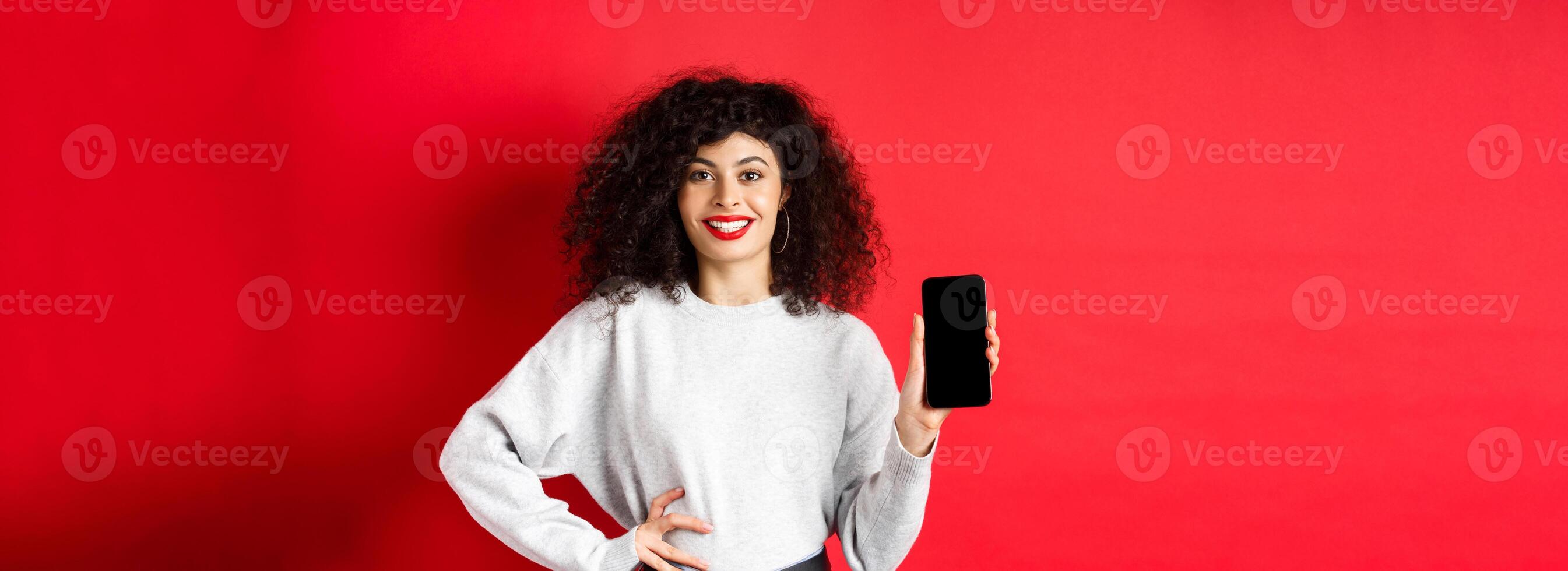 Attractive smiling woman showing empty smartphone screen and looking happy, advertising online store or application, standing against red background photo