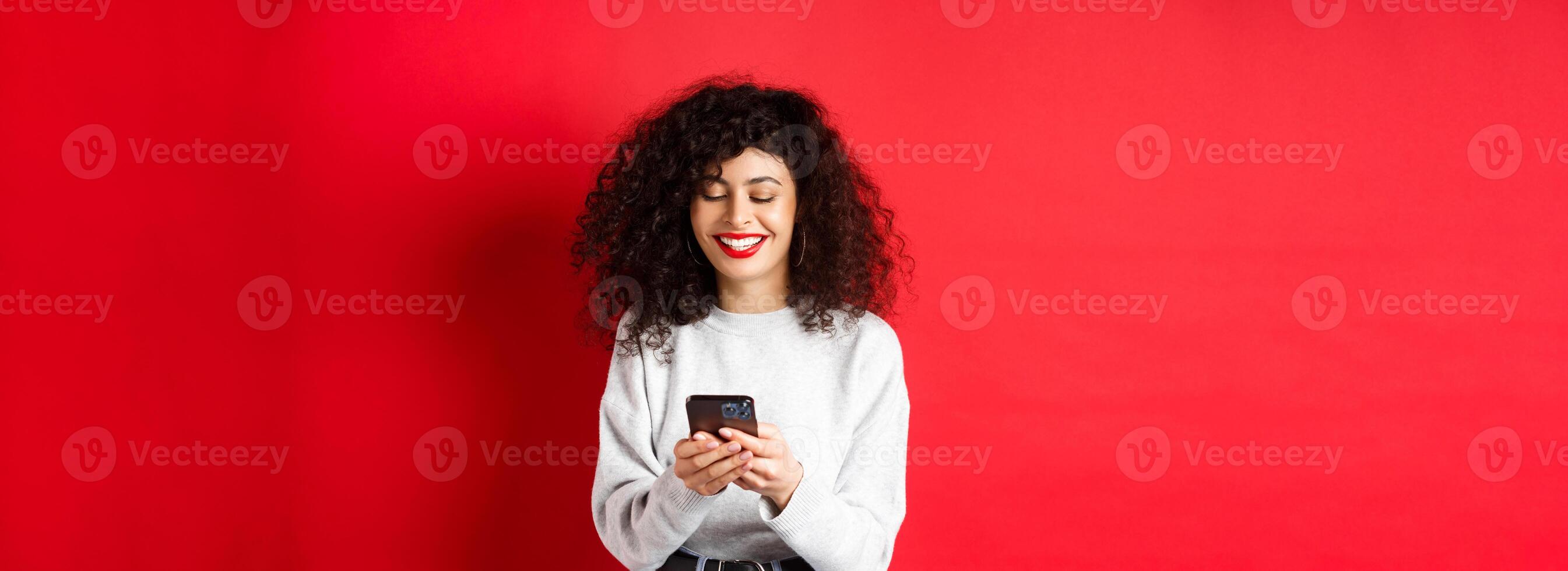 Happy positive girl chatting on phone, reading message and smiling, using social media app, standing on red background photo