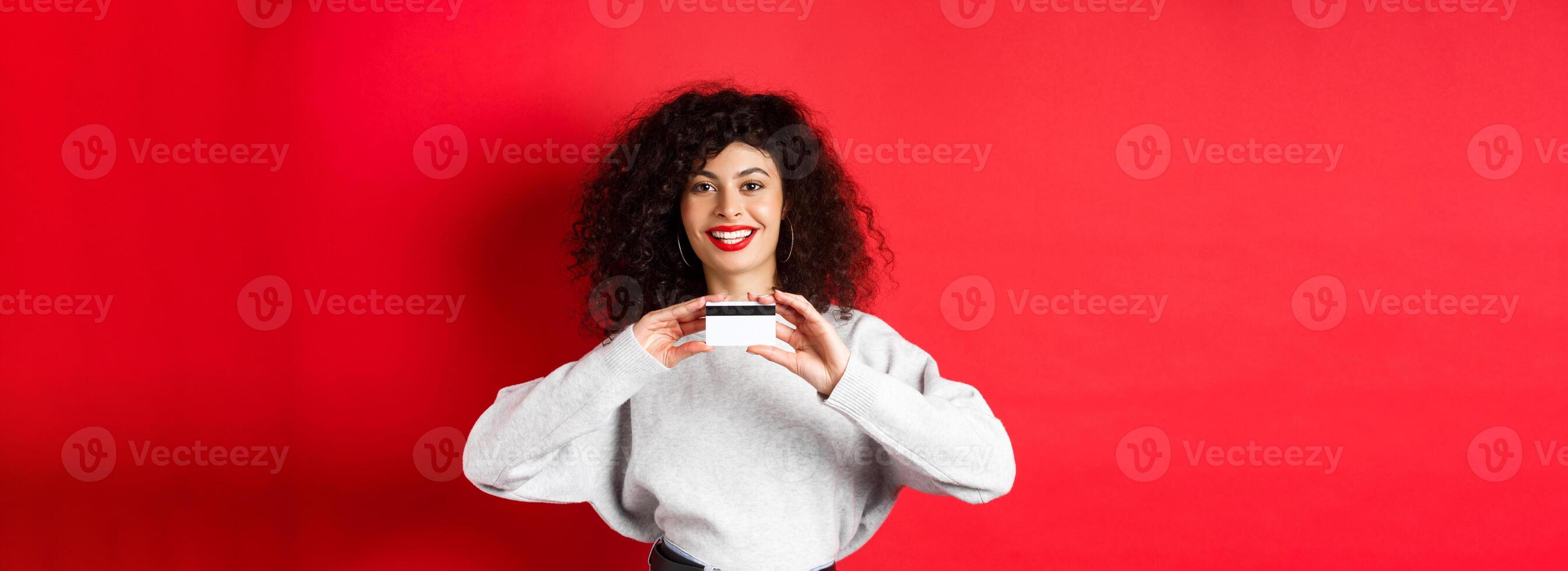 Happy lady with curly hair showing plastic credit card and smiling, standing against red background. Shopping concept photo