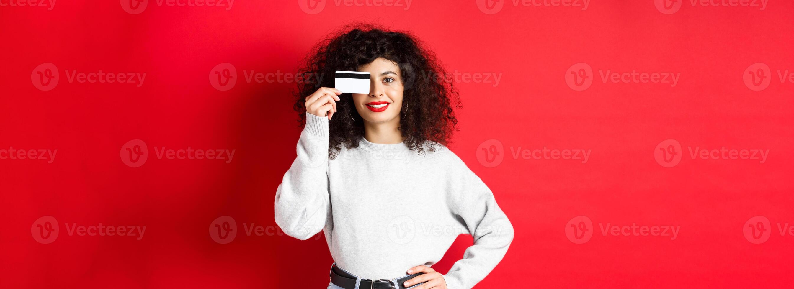Beautiful female model with curly hair, showing plastic credit card and smiling, standing on red background. Makeup and shopping concept photo