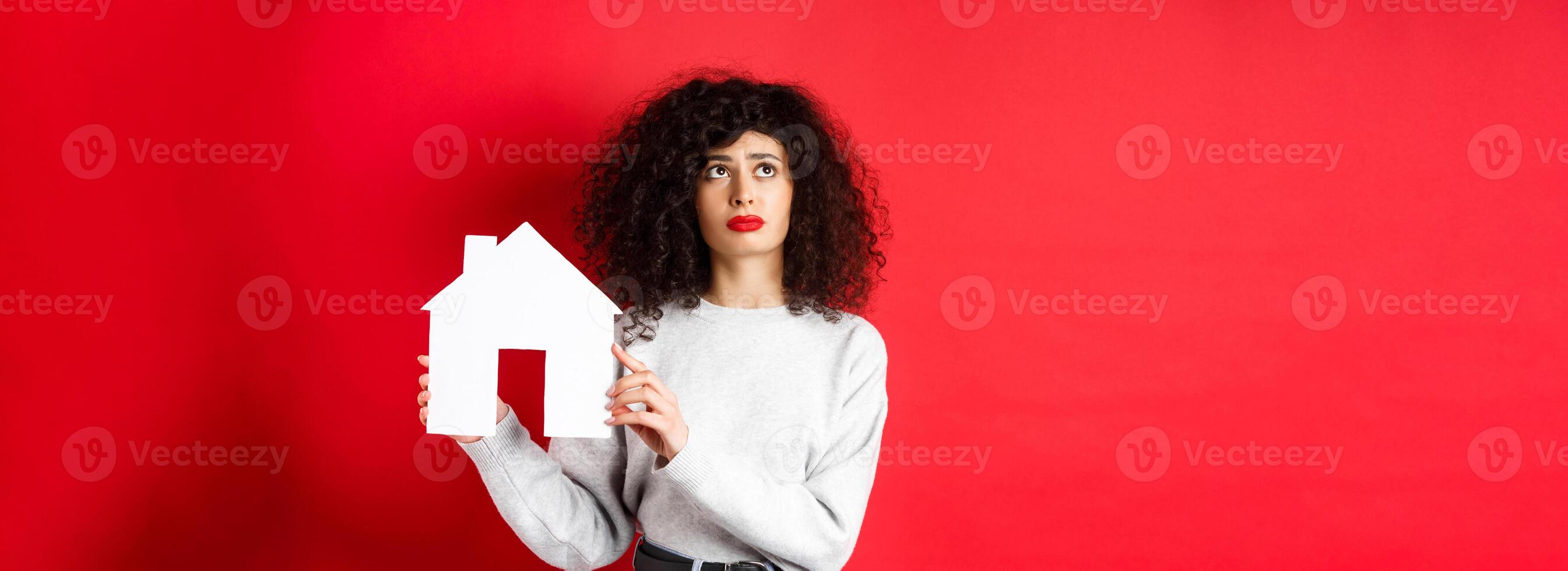Real estate. Sad woman thinking of buying own house, showing paper home cutout and looking up with uspet face, standing on red background photo