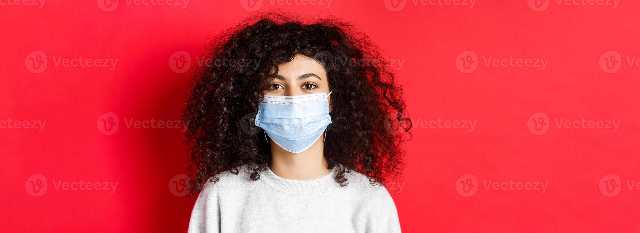Covid-19 and pandemic concept. Close-up of modern young woman with curly hair, wearing medical mask from coronavirus, smiling at camera, red background photo