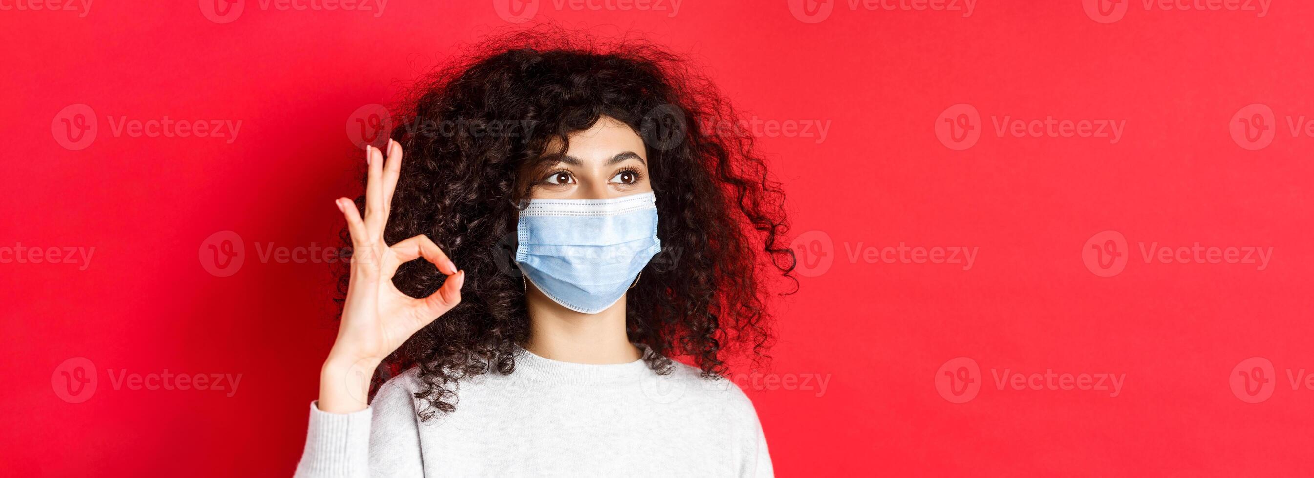 Covid and health concept. Image of young woman in medical mask approve promo, showing okay gesture and looking at empty space, standing on red background photo