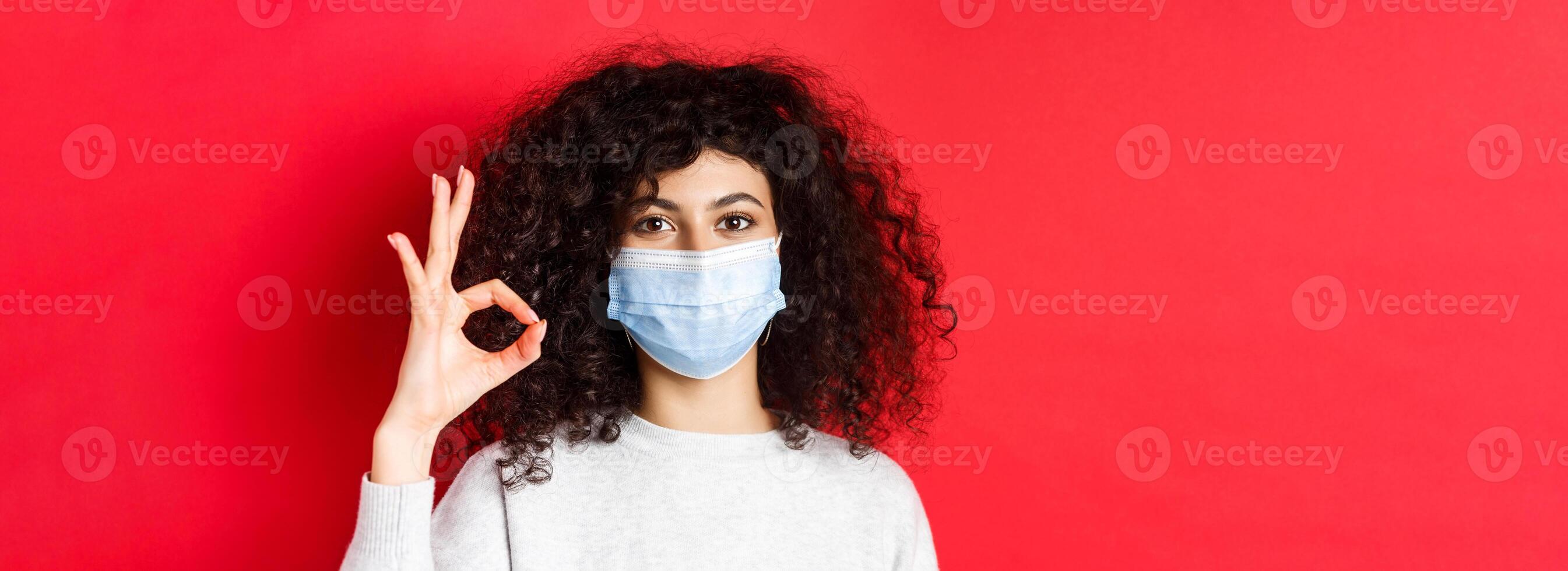 Covid and health concept. Confident young woman in medical mask showing OK gesture in approval, guarantee quality, standing on red background photo