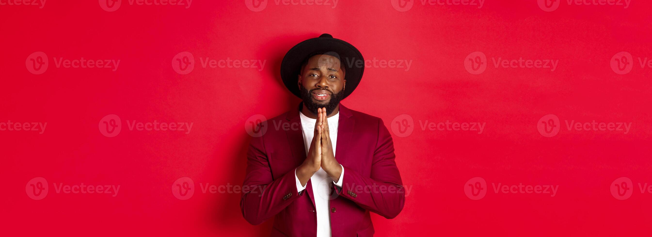Miserable Black man asking for help, begging for favour and looking sad, standing against red background photo