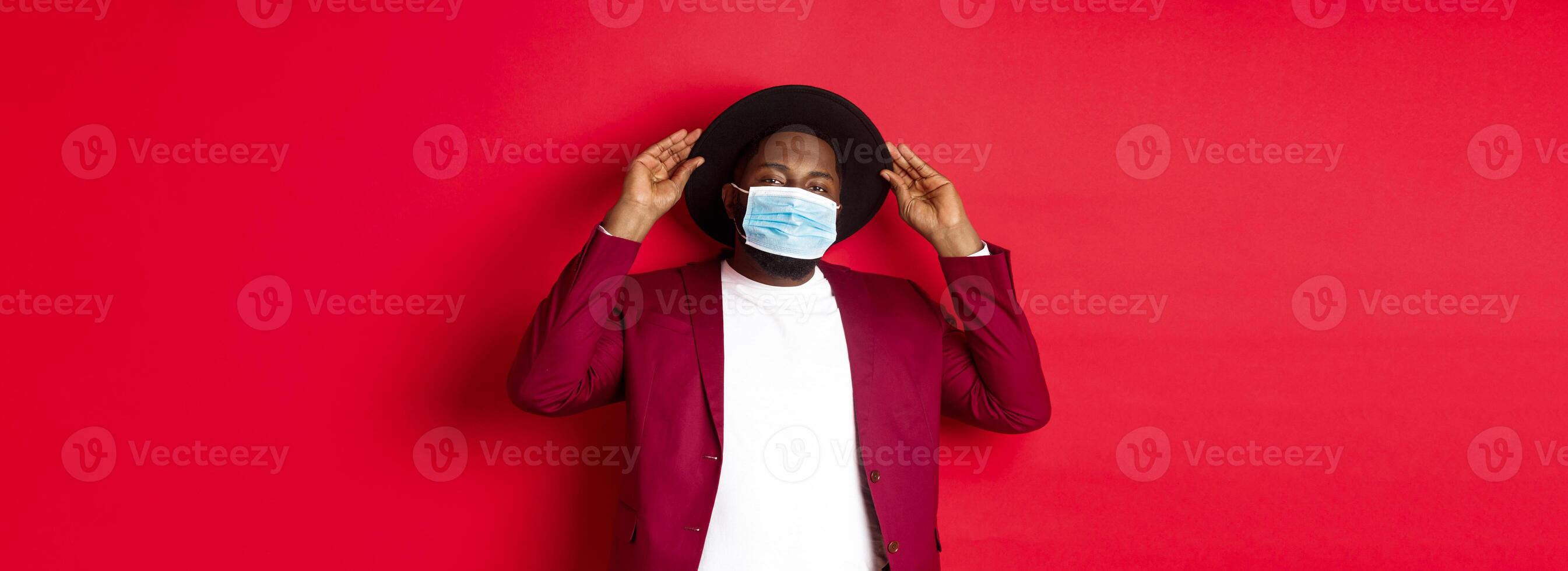 Covid-19, quarantine and holidays concept. Handsome and stylish african american man in face mask, put hat on head and looking sassy, dress-up for party, standing over red background photo