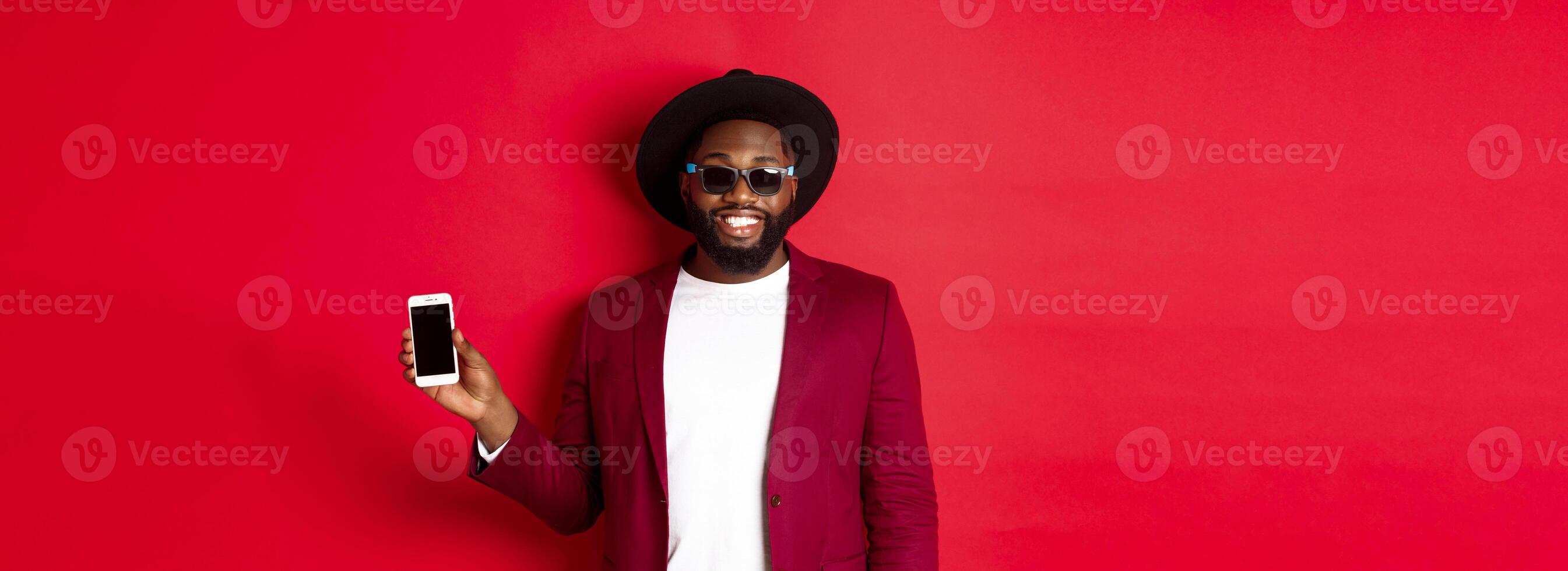 Handsome and stylish Black man showing phone screen at camera, recommending online store or application, standing over red background photo