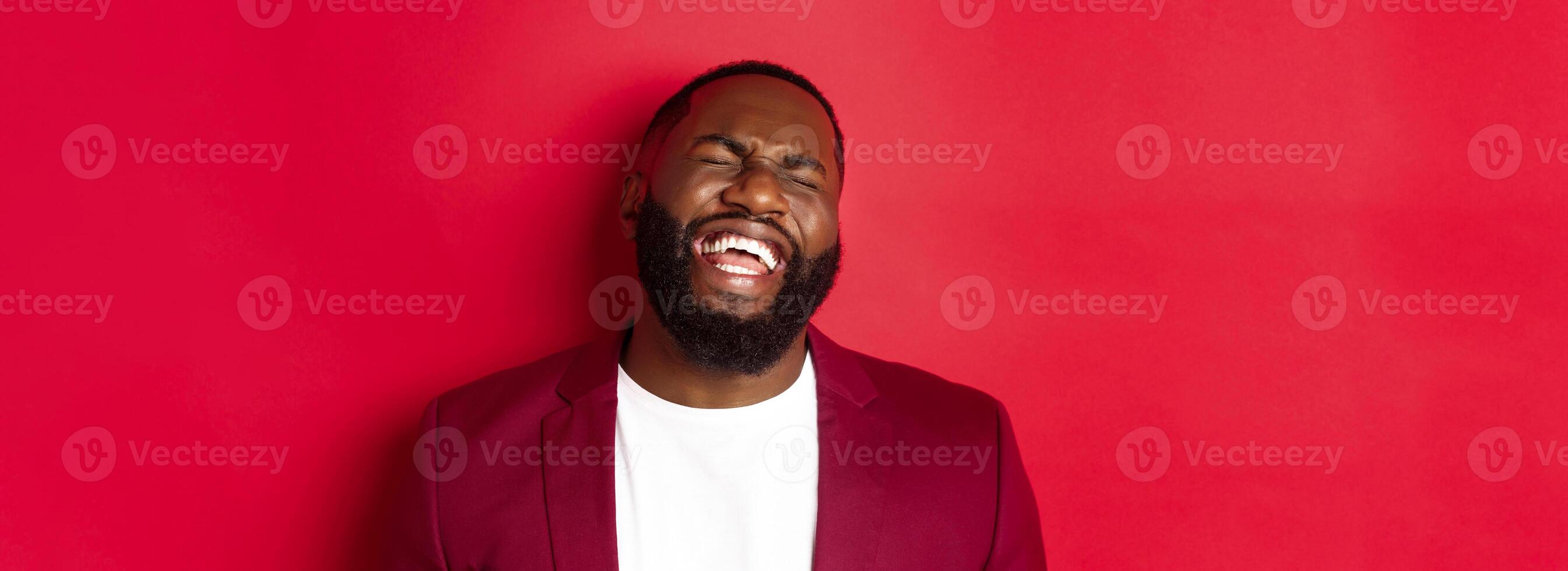 Close-up of happy and carefree Black man having fun, laughing and smiling, standing in blazer against red background photo