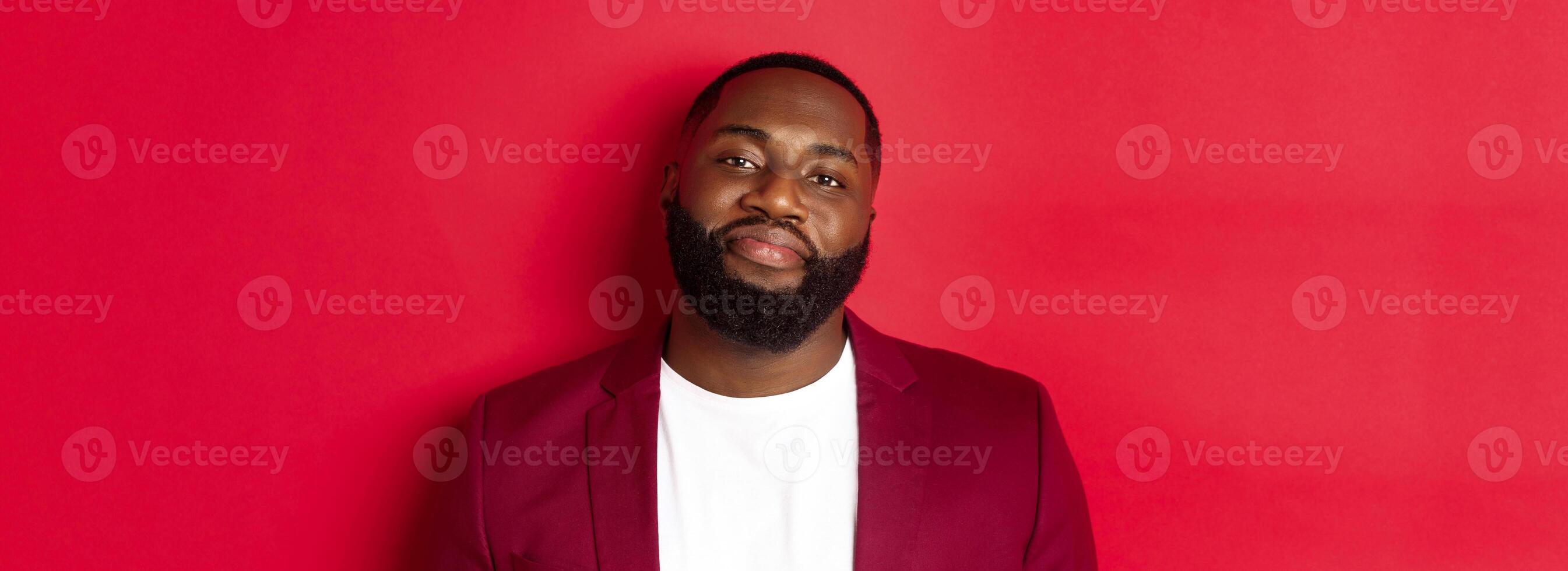 Close-up of handsome bearded plump guy smiling at camera shy, standing in stylish blazer on t-shirt, standing over red background photo