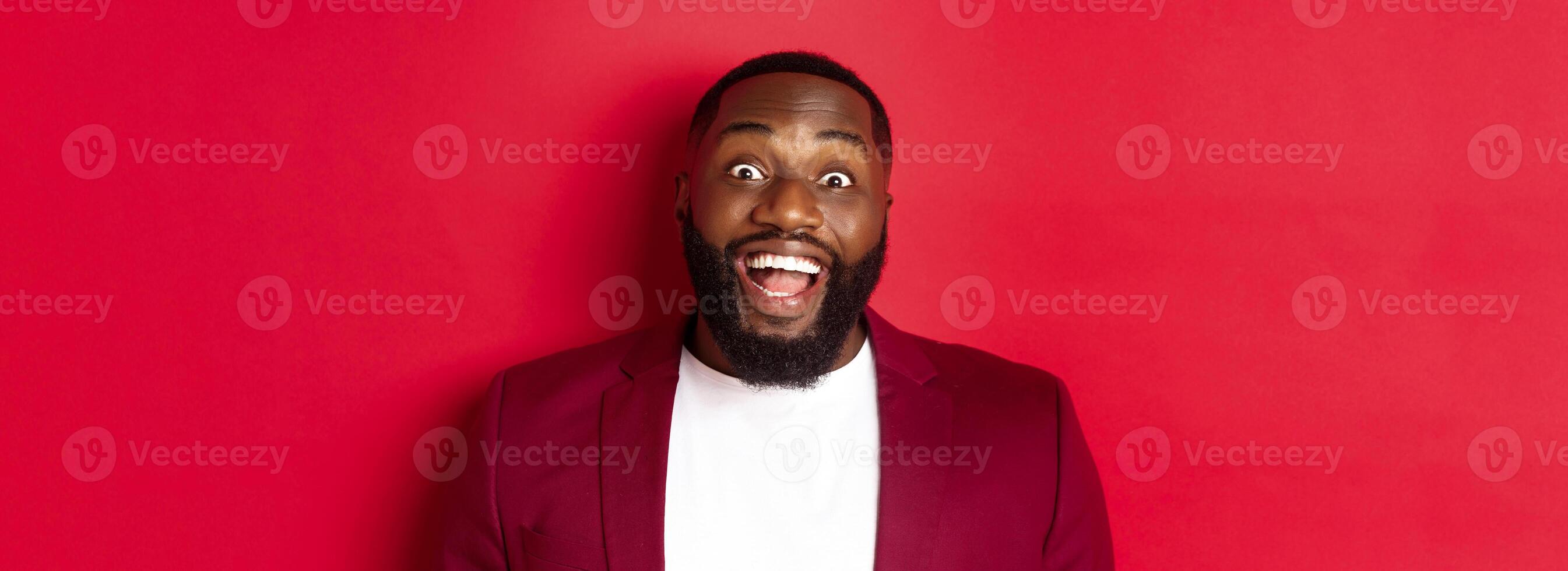 Close-up of african american man with beard staring amazed at camera, smiling and looking excited, standing in blazer against red background photo