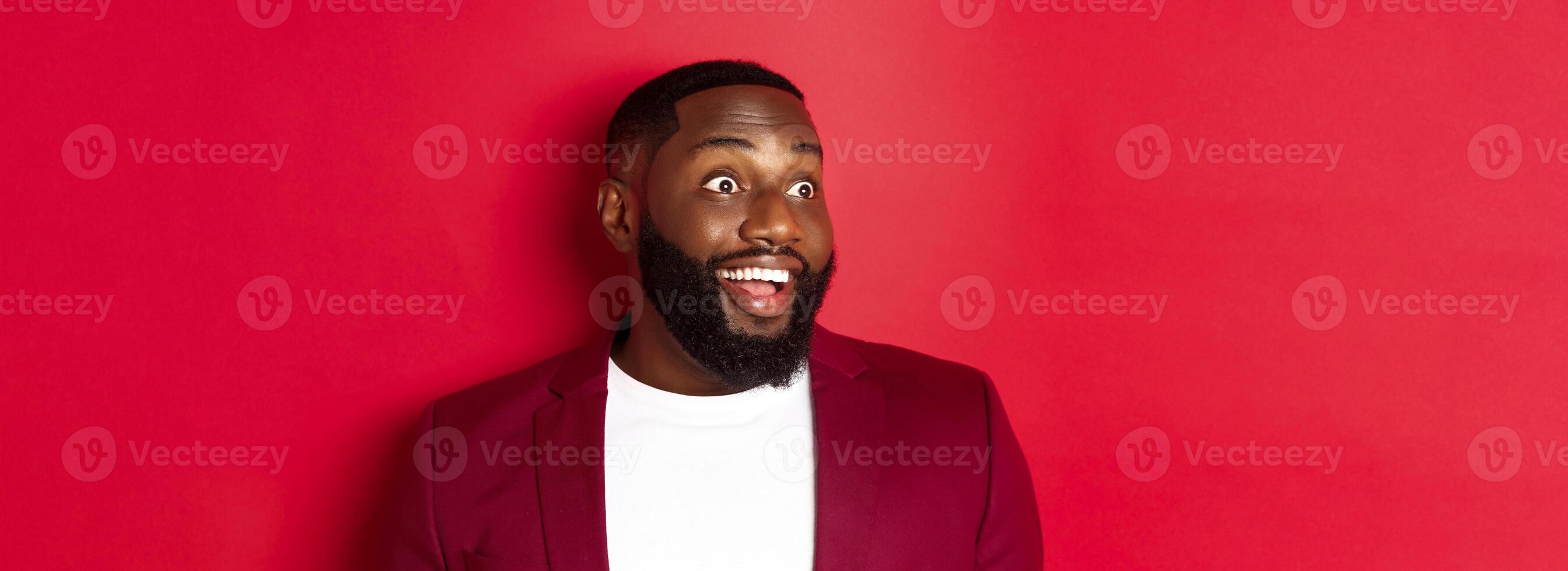 Close-up of excited african american guy looking left, smiling with amazement and joy, standing in blazer against red background photo