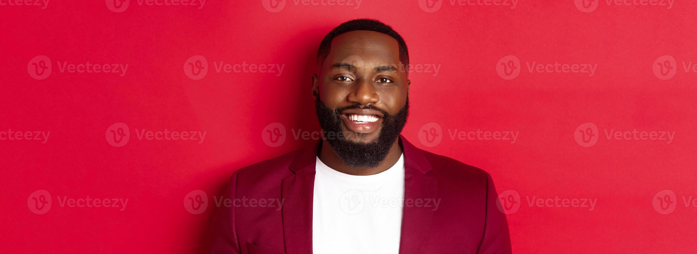 Close-up of handsome and stylish Black man wearing party blazer, smiling happy at camera, standing against red background photo