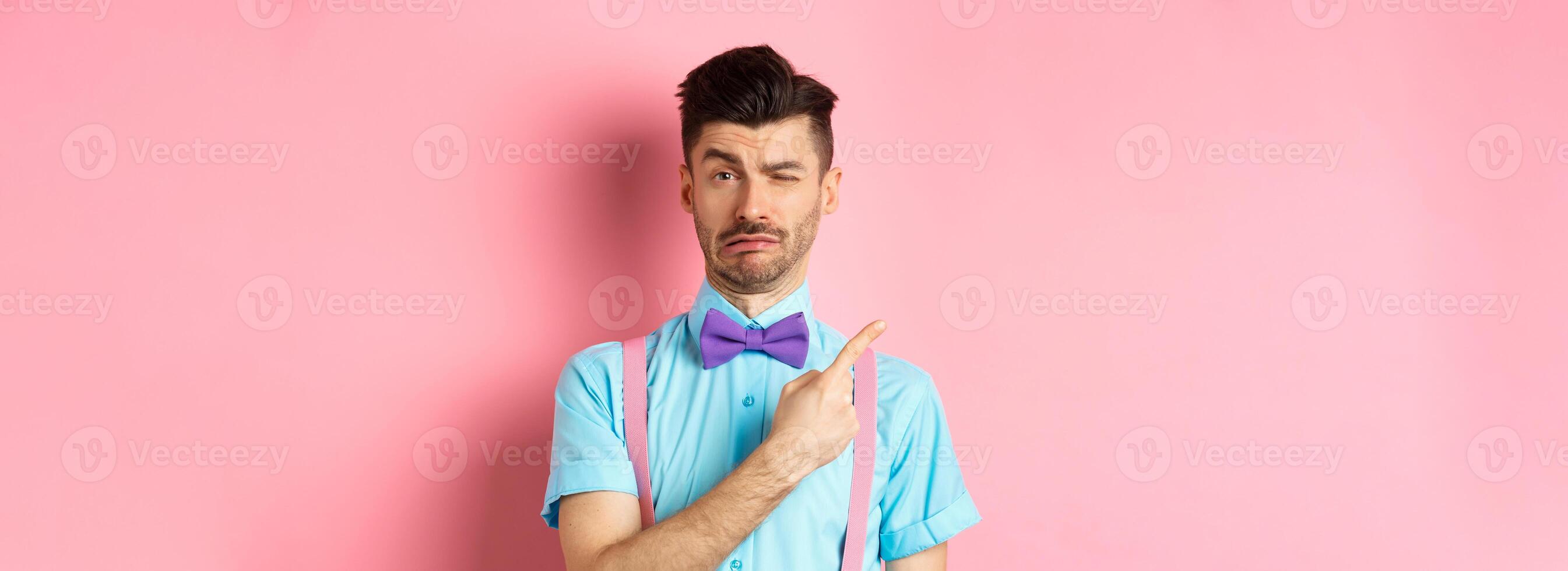 Childish young man acting sad and fake crying while pointing finger upper left corner, complaining on something, standing over pink background photo
