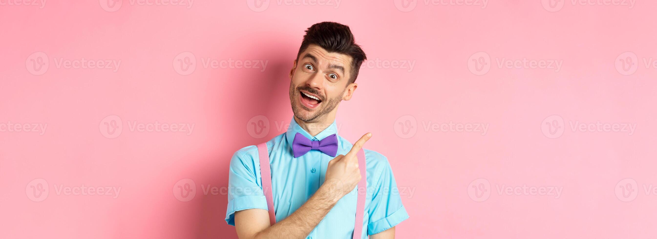 Funny male model in bow-tie, tilt head and pointing finger left, inviting to look, check it out gesture, showing advertisement on pink background photo