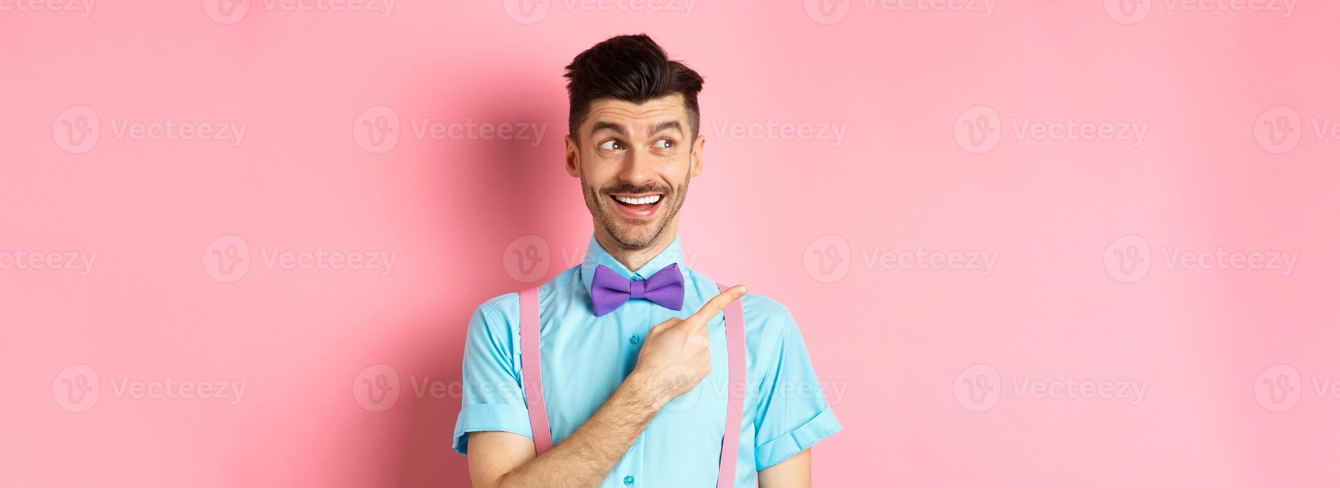 Cheerful caucasian man in funny bowtie looking and pointing hand left, showing promo logo, standing on pink background photo