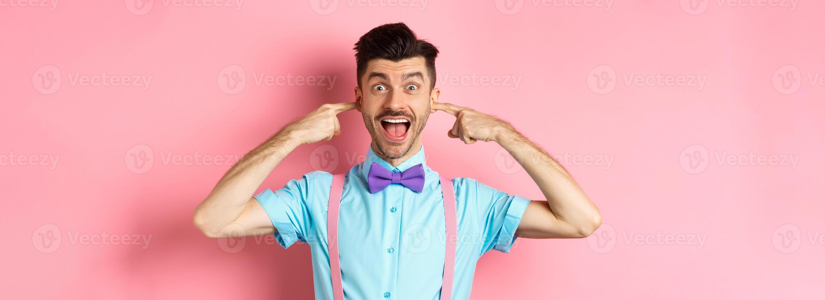 Childish funny guy shut ears and laughing at camera, refuse to listen, standing ignorant on pink background photo
