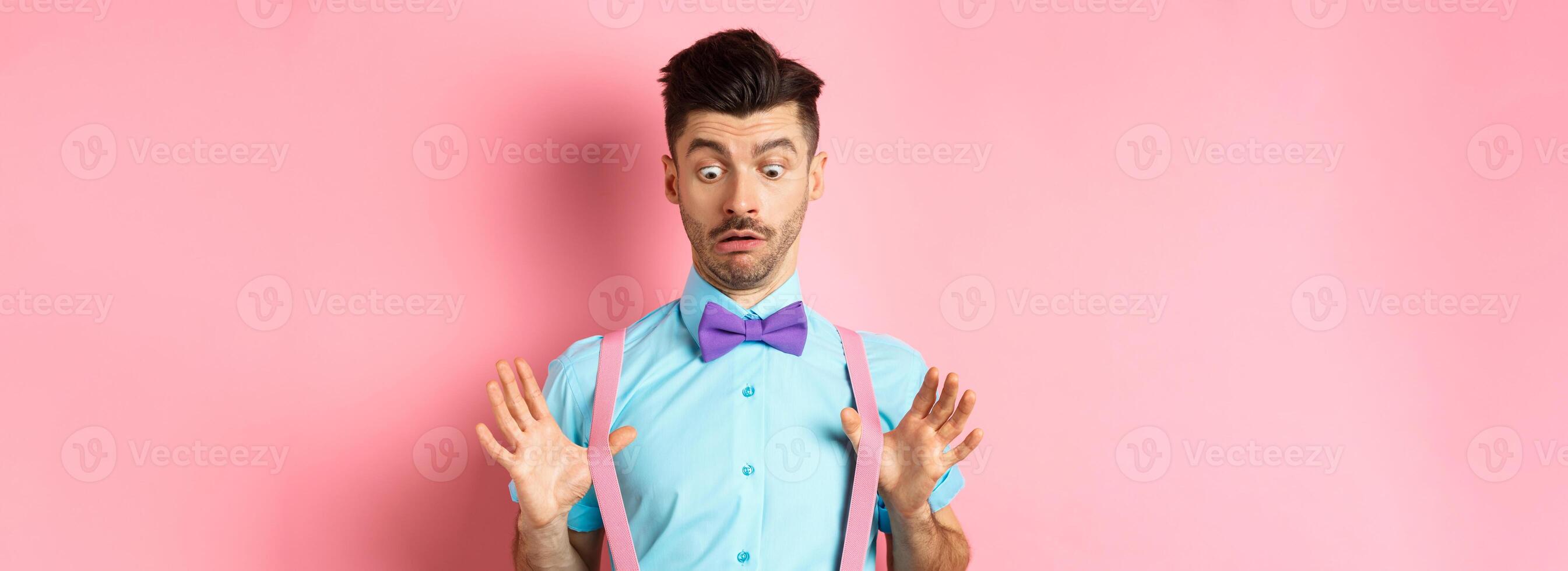 Funny guy with moustache and bow-tie, adjusting his suspenders and looking down with confused and surprised face, standing over pink background photo