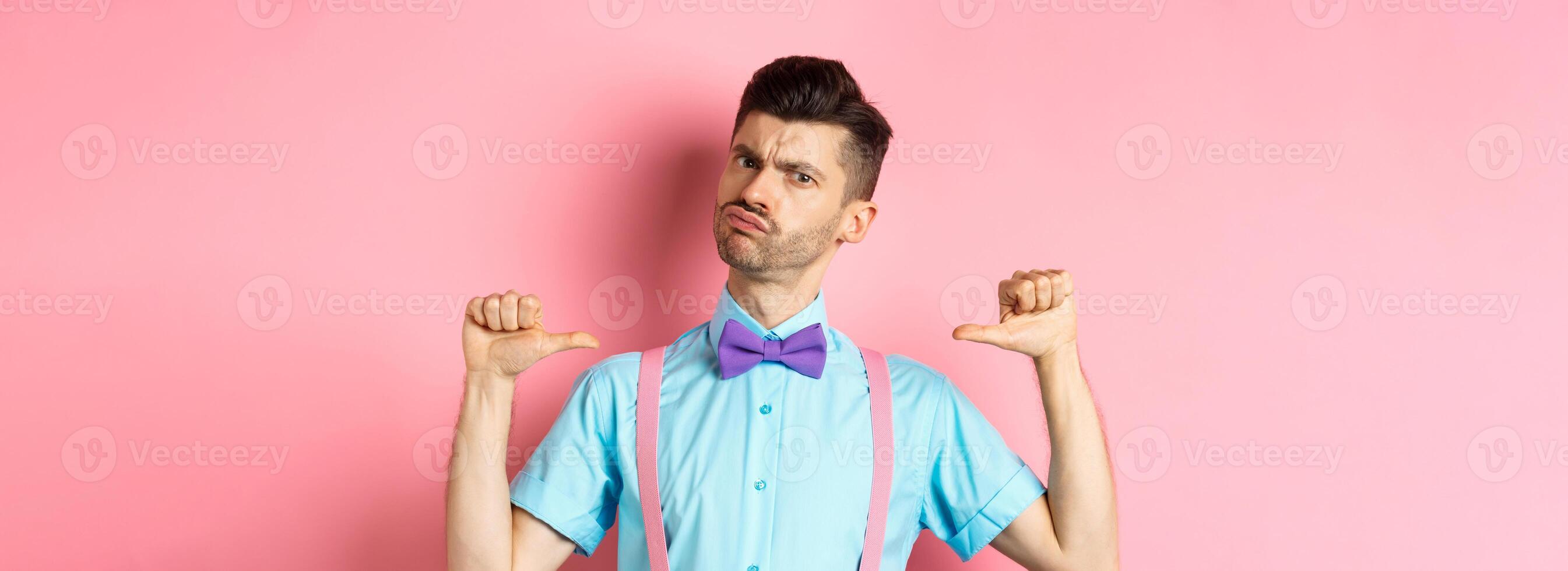 Serious and funny young man trying look cool, pointing at himself to self-promote, being a professional, standing over pink background photo