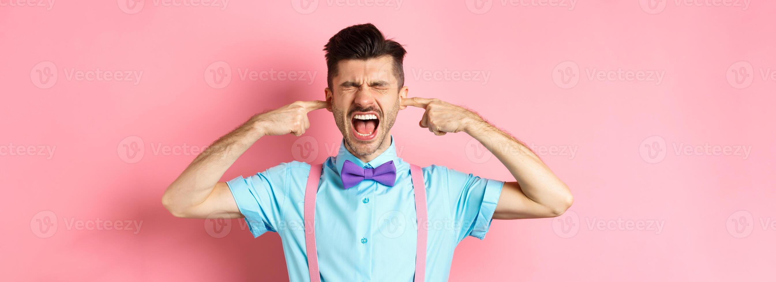 Irritated young man block noise, shut ears with hands and screaming annoyed, being disturbed by loud sound, standing over pink background photo