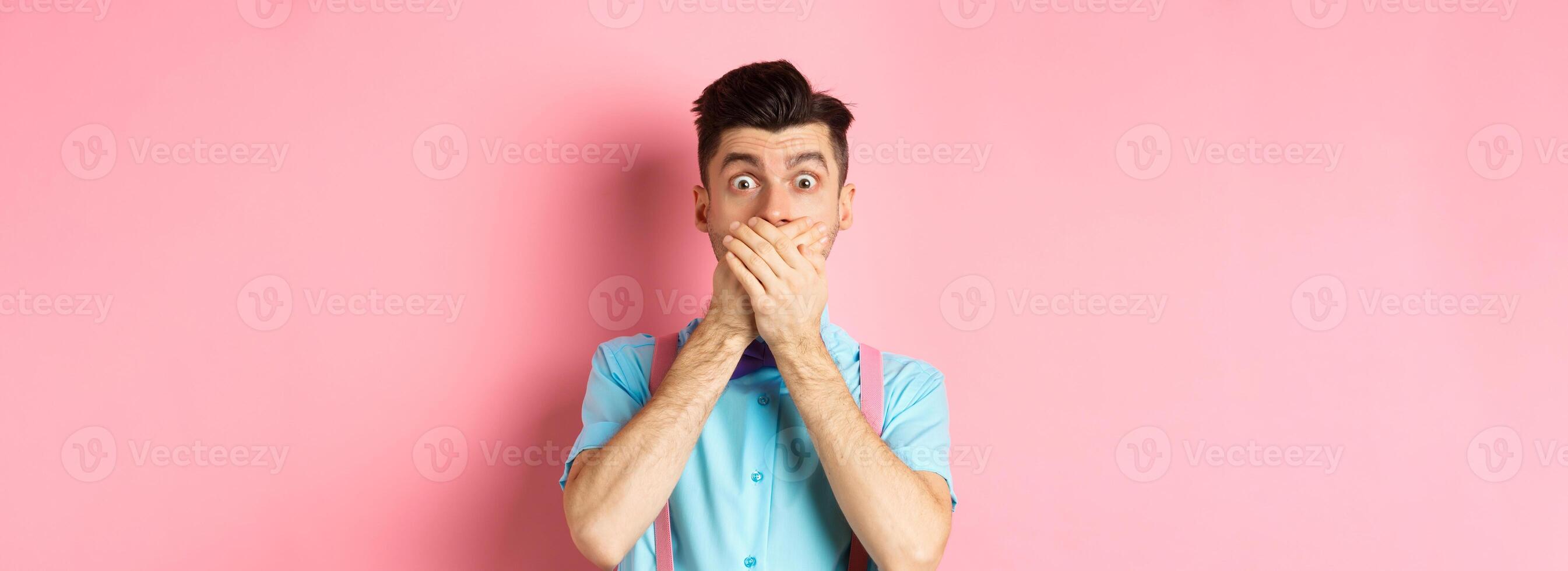 Shocked and speechless guy shut his mouth, cover lips with hands and staring startled at camera, standing over pink background photo