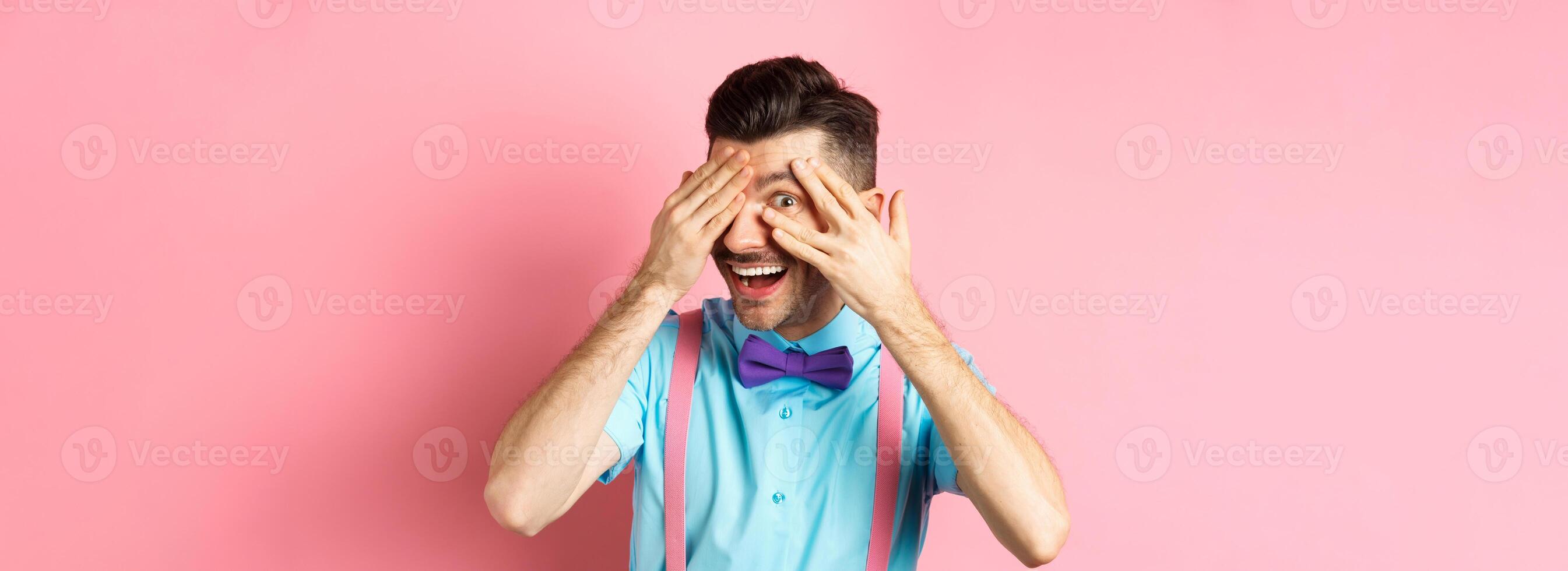 Intrigued funny guy in bow-tie, peeking through fingers at something interesting, checking out something cool, standing on pink background photo