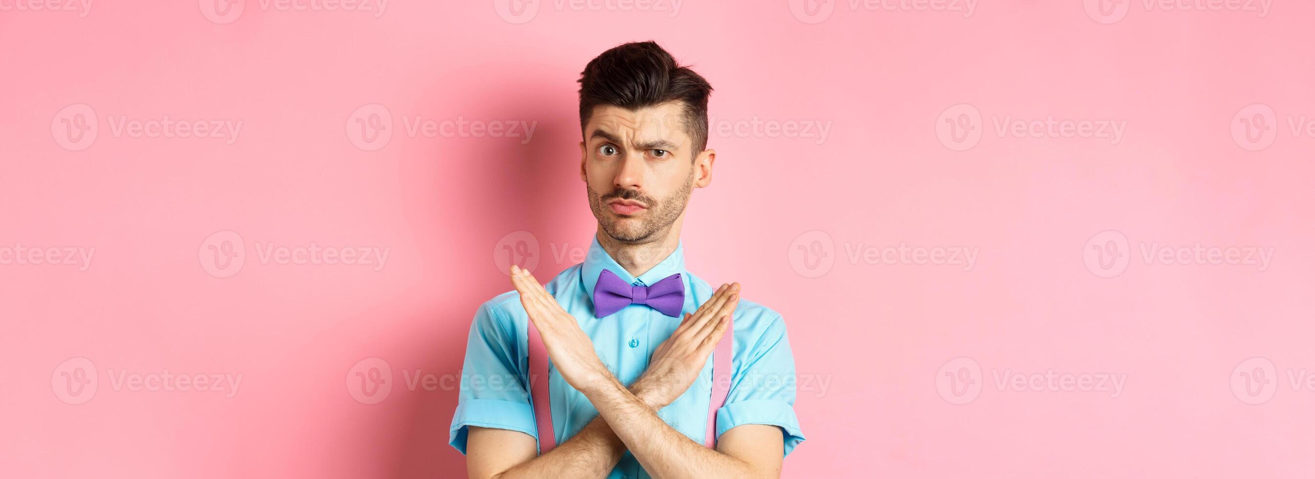 Serious-looking skeptical guy showing cross sign to stop, prohibit something bad, forbid action, standing on pink background photo