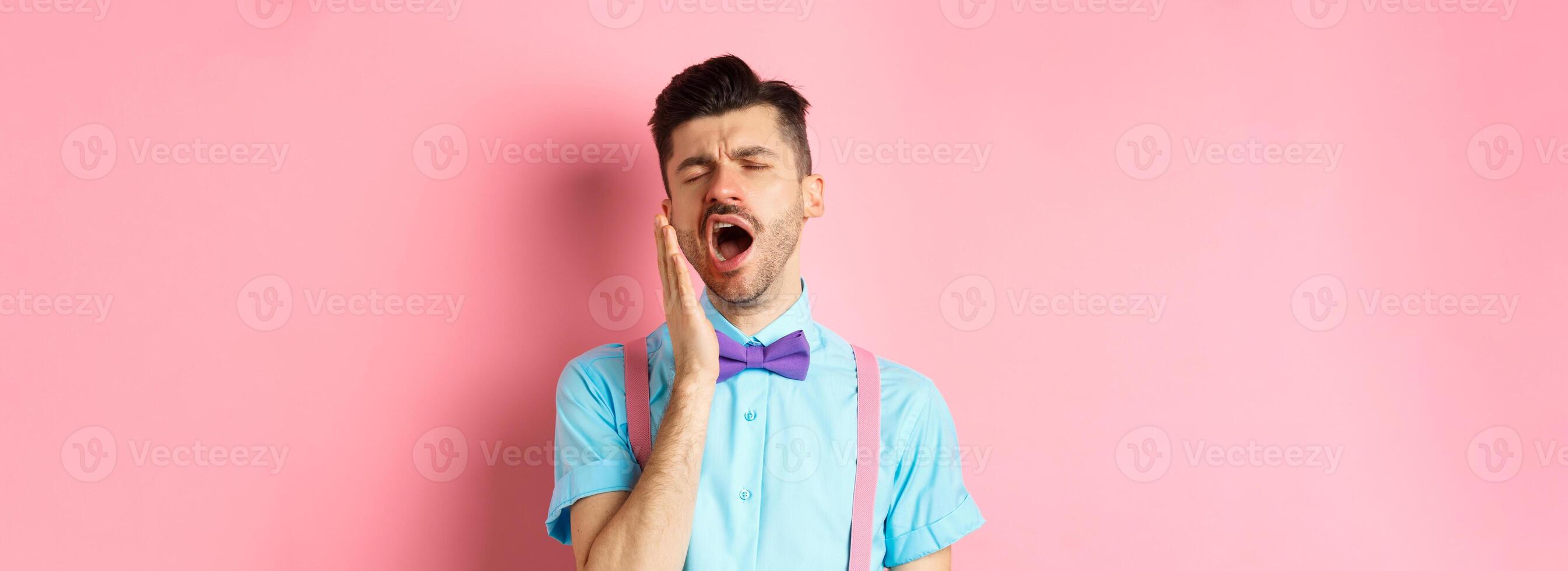 Image of tired guy in bow-tie yawning with opened mouth and closed eyes, standing sleepy, feeling fatigue, standing over pink background photo