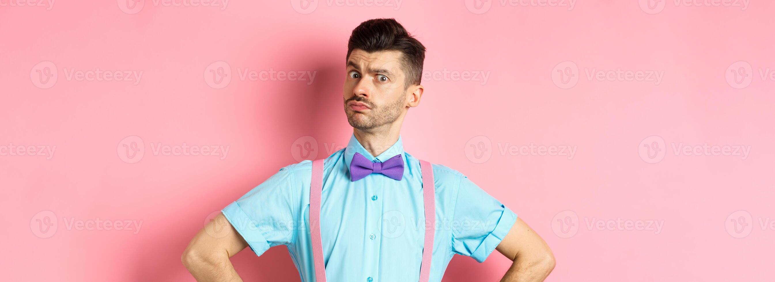 Image of young man with funny moustache and bow-tie standing in confident pose, staring with disbelief and doubts at camera, posing over pink background photo
