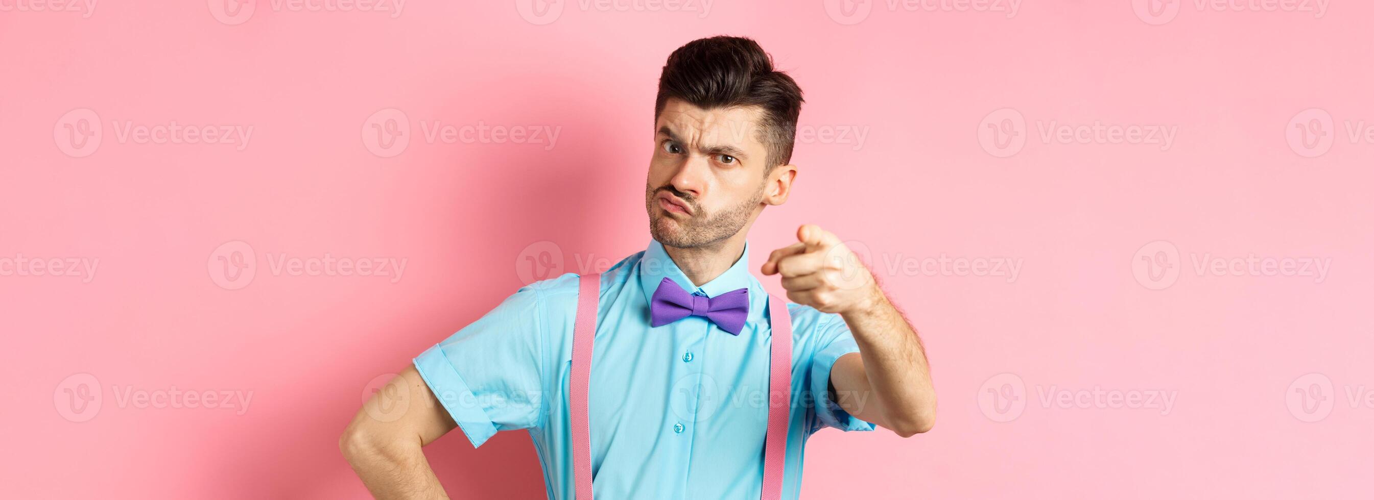 Grumpy guy in bow-tie and suspenders accusing you, its your fault gesture, pointing finger at camera with disappointed frowning face, pink background photo