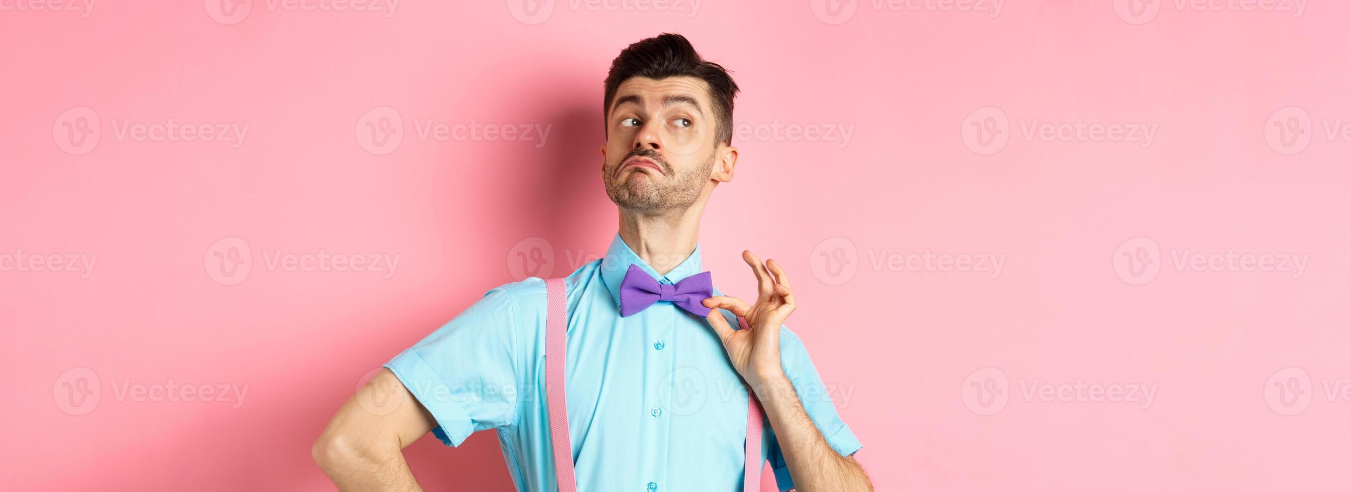 Image of funny man raising chin up and adjusting bow-tie, standing confident and sassy on pink background, looking left photo