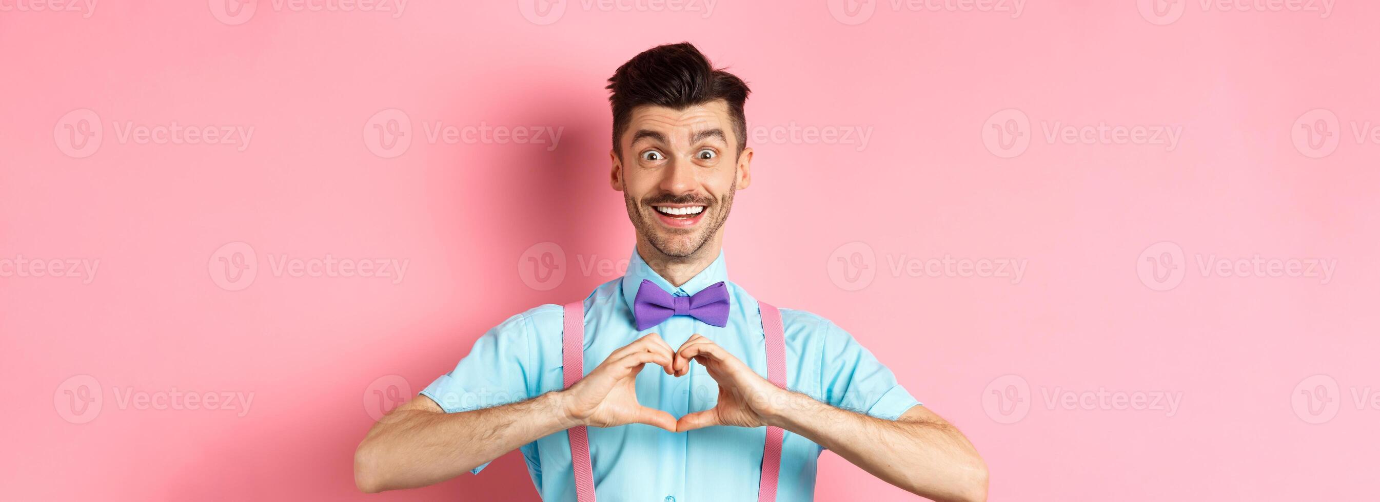 Happy Valentines day. Smiling funny guy with moustache showing heart sign and looking at lover, saying I love you, express feeling, standing over romantic pink background photo