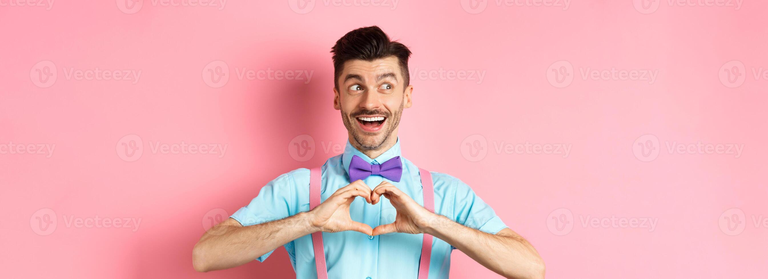 Happy Valentines day. Cheerful male model enjoying romantic date, showing heart sign, say I love you and looking left with happy smile, standing over pink background photo