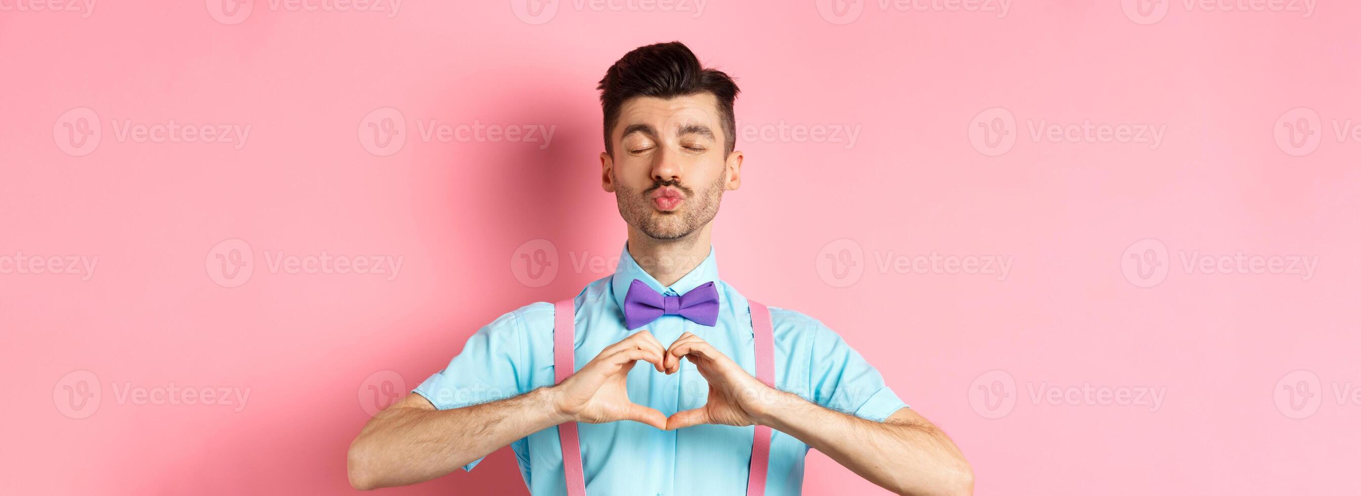 Happy Valentines day. Romantic boyfriend close eyes for kiss and showing heart, having date with lover, standing over pink background photo