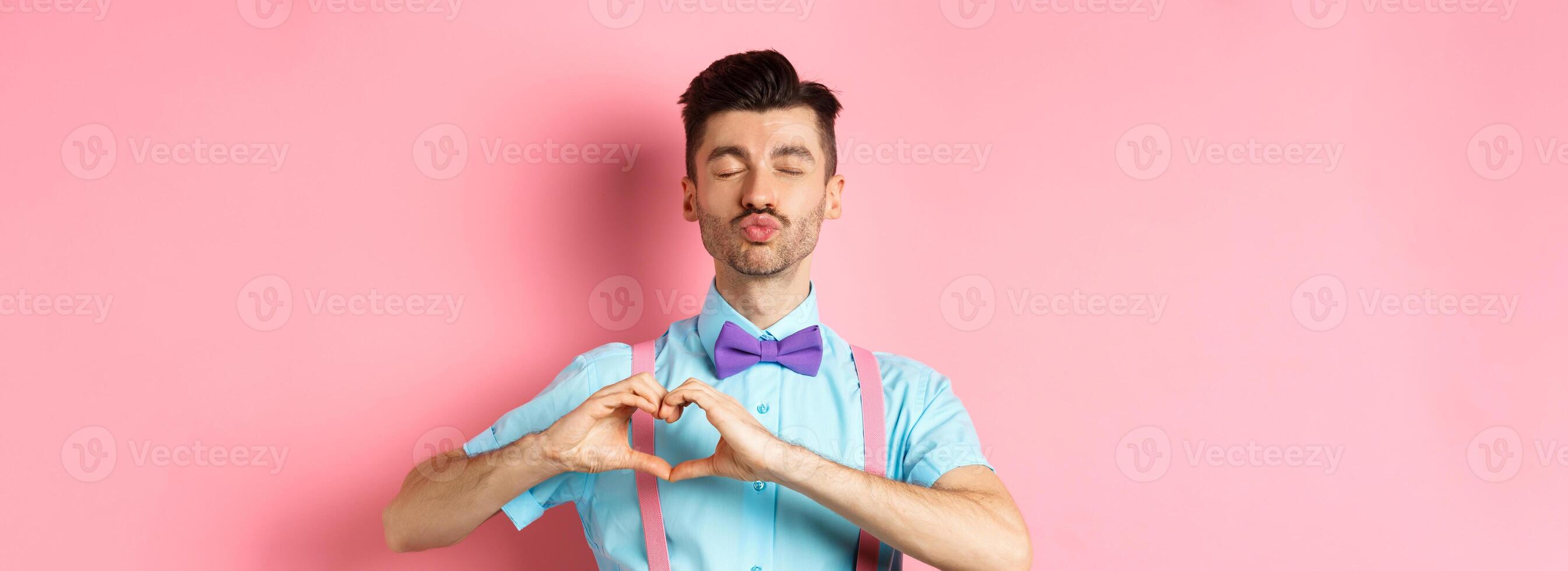 Romantic man in funny bow-tie waiting for kiss, close eyes and pucker lips kissing, showing heart gesture, being in love on Valentines day, standing over pink background photo