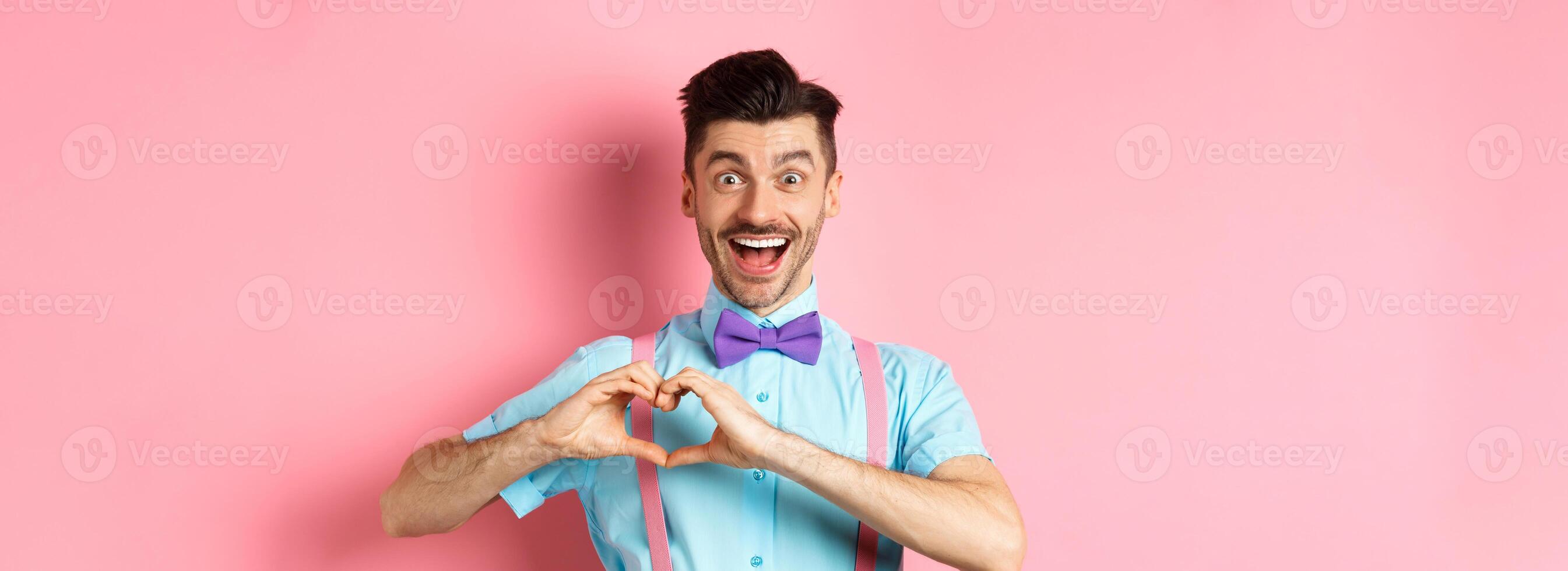 Excited smiling man showing pounding heart and looking with love, standing over romantic pink background. Valentines day concept photo