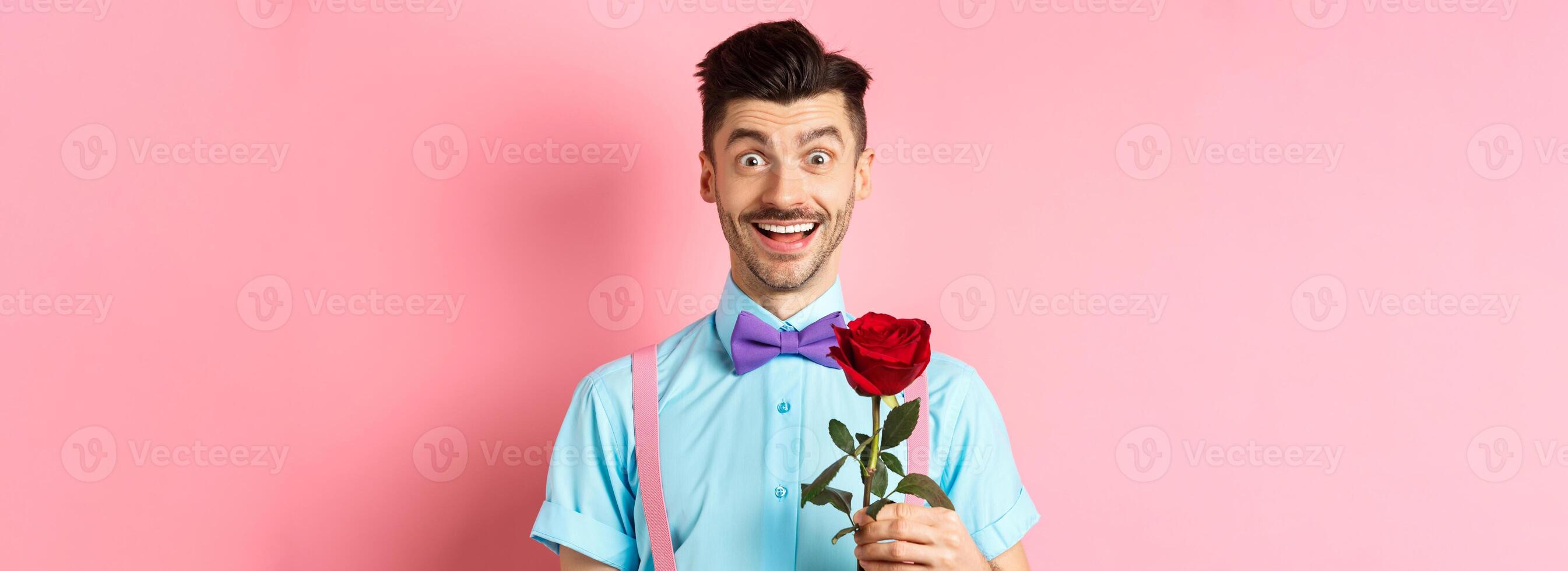 Excited bearded man with moustache and bow-tie waiting for date with red rose, having romantic moment on Valentines day, standing on pink background photo