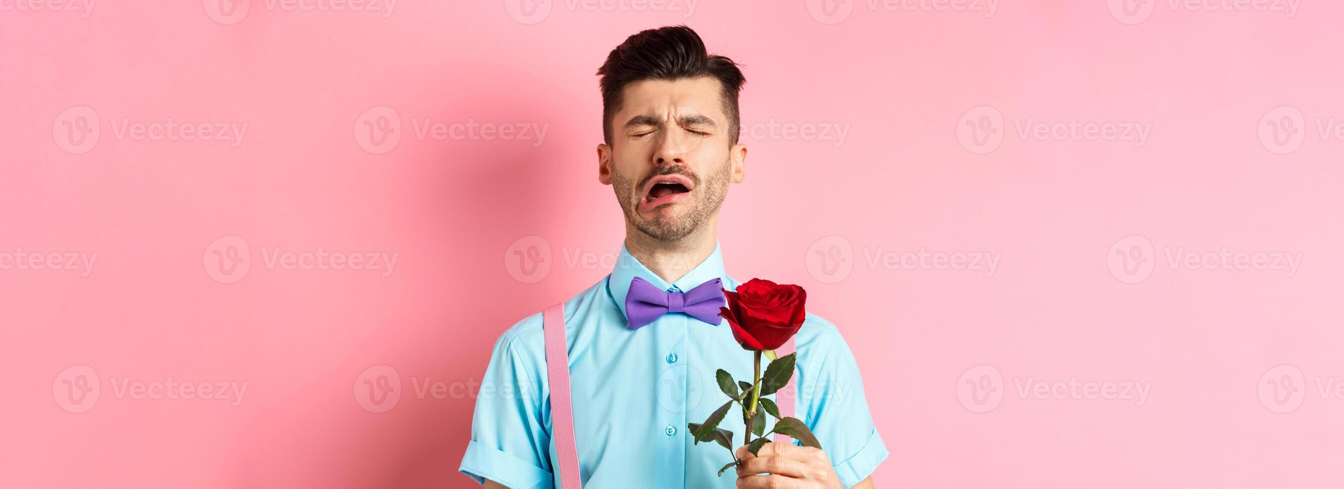 Heartbroken guy in funny bow-tie crying over girlfriend, standing alone with red rose on pink background and sobbing, break-up on Valentines day photo