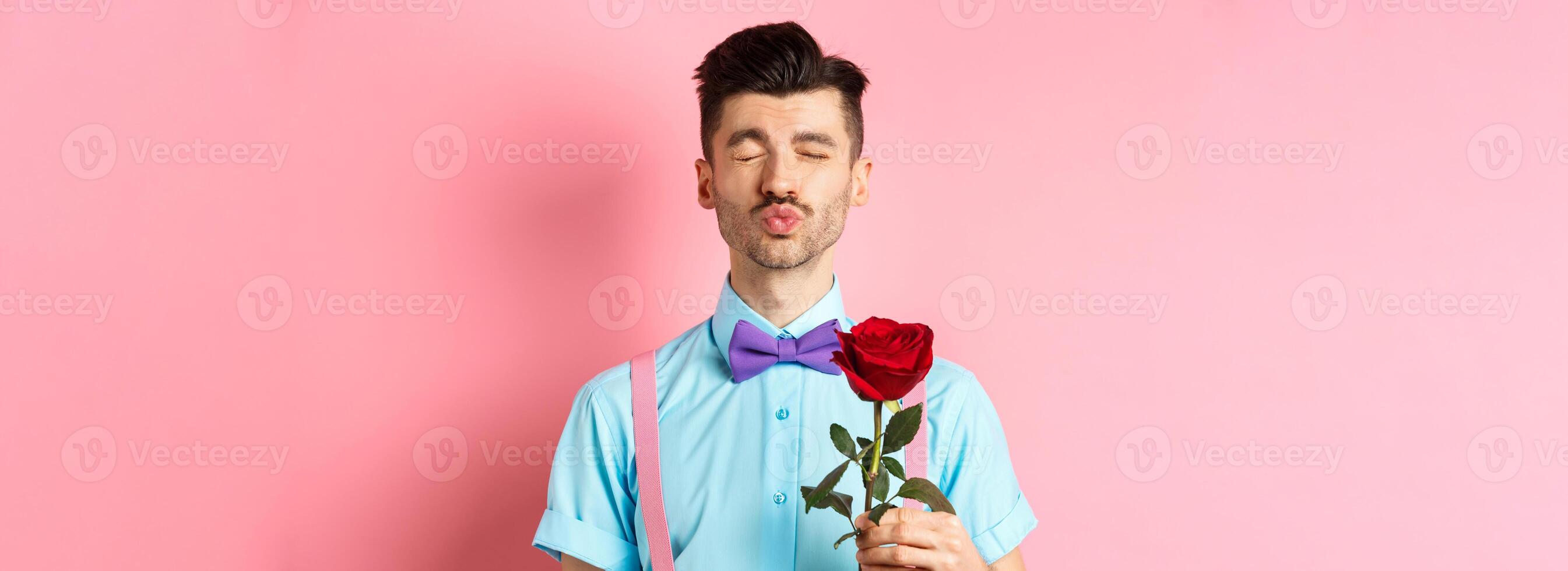 Cute and funny man waiting for kiss from lover on Valentines day, holding beautiful red rose for girlfriend, standing over pink background photo