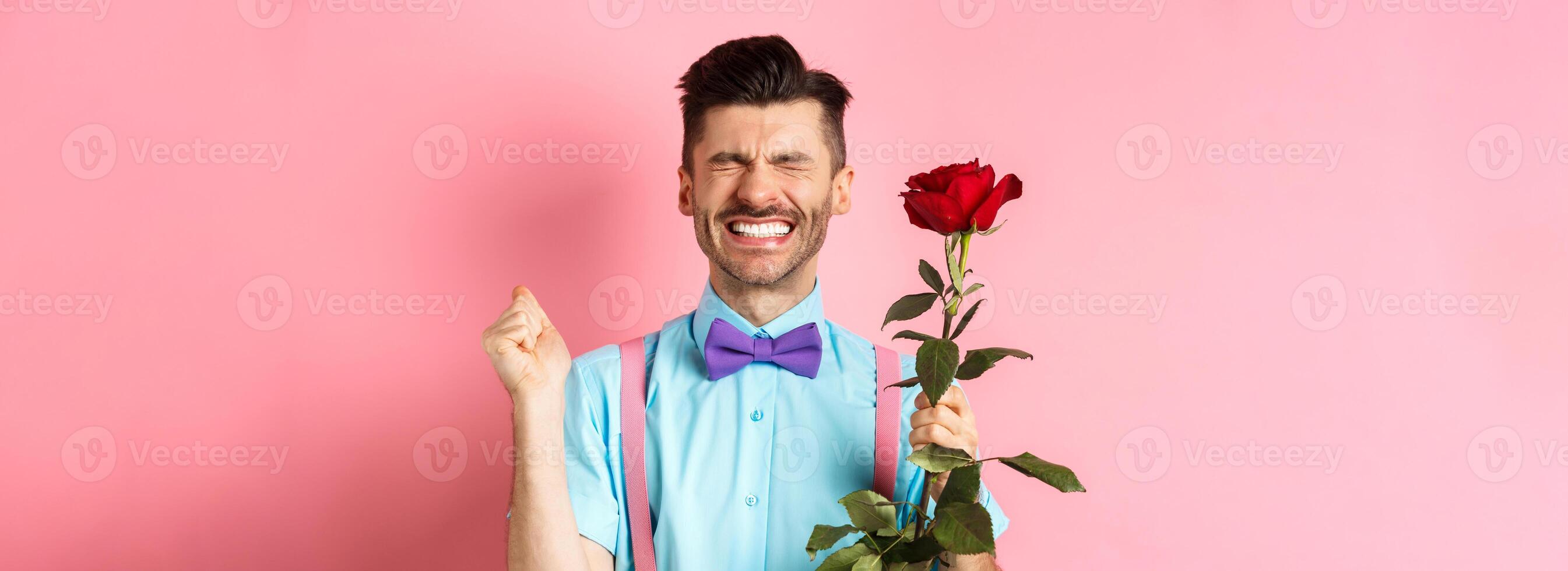 romance y san valentin día concepto. esperanzado hombre sensación emocionado antes de fecha, esperando para amante con rojo Rosa y Orando, en pie terminado rosado antecedentes foto
