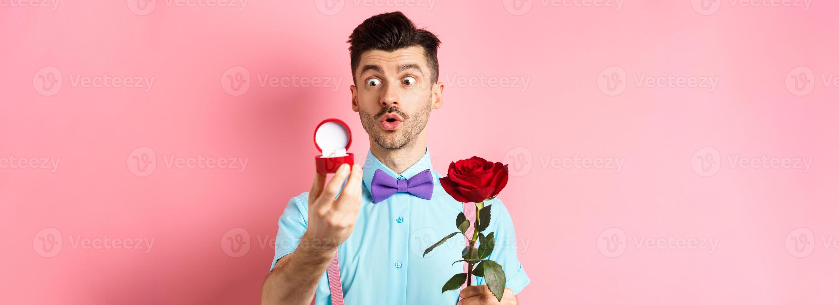 Valentines day. Romantic funny guy going to make wedding proposal, asking to marry him, holding red rose and looking at engagement ring with excitement, pink background photo